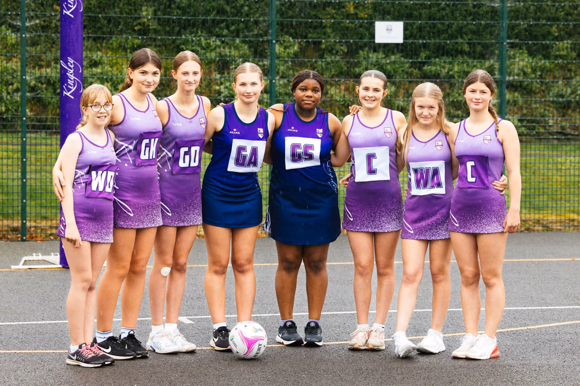 Eight young female netball players standing side by side on an outdoor court, wearing purple and navy team uniforms with position letters, smiling at the camera with a netball on the ground in front of them.