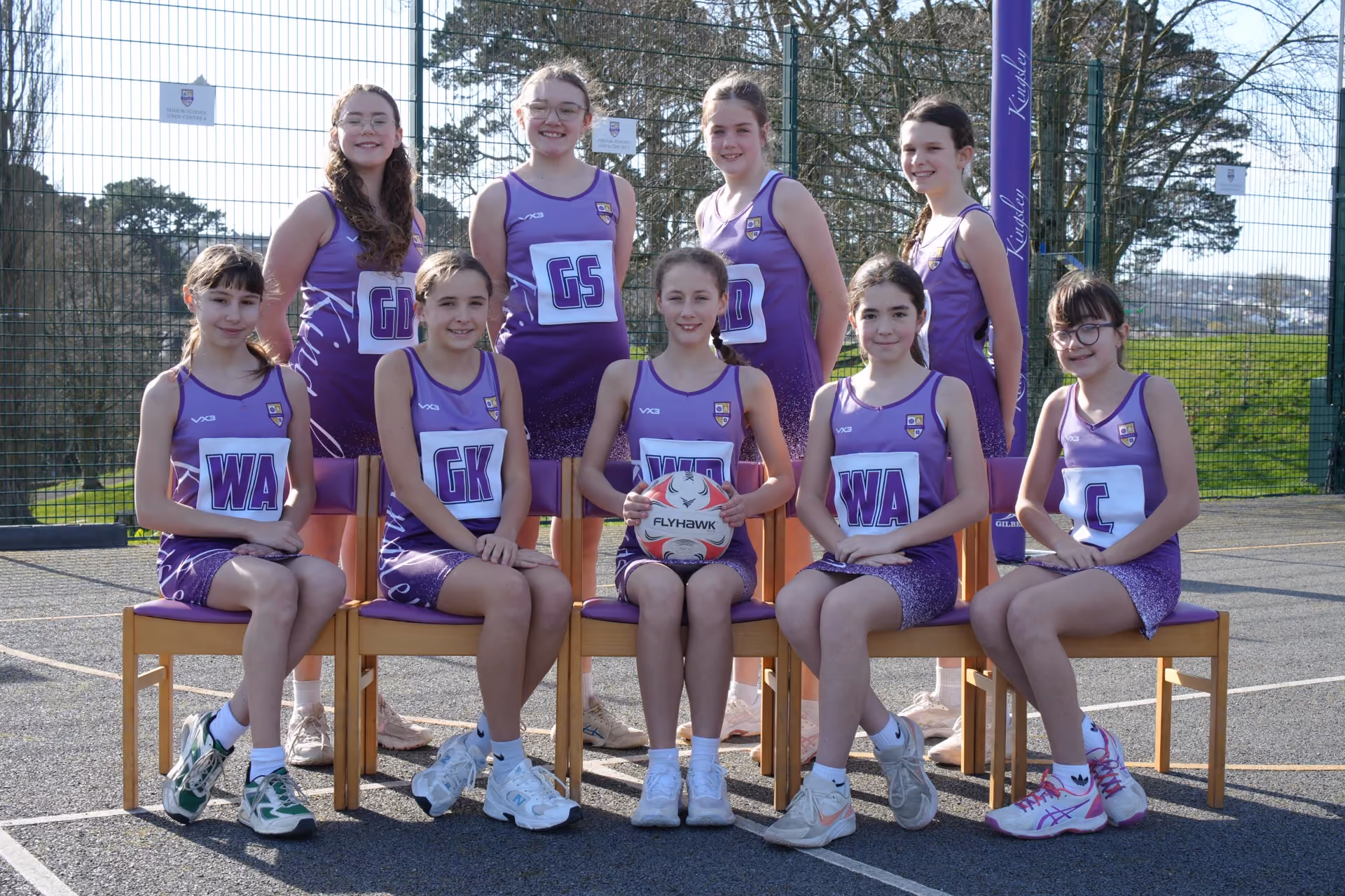 A group of nine girls in purple netball uniforms posing outdoors on a netball court with a netball stuck between their legs in the front center. Five girls are seated on chairs in the front row, and four girls are standing behind them. Their uniforms display netball position abbreviations like WA, GK, GS, GD, and C.