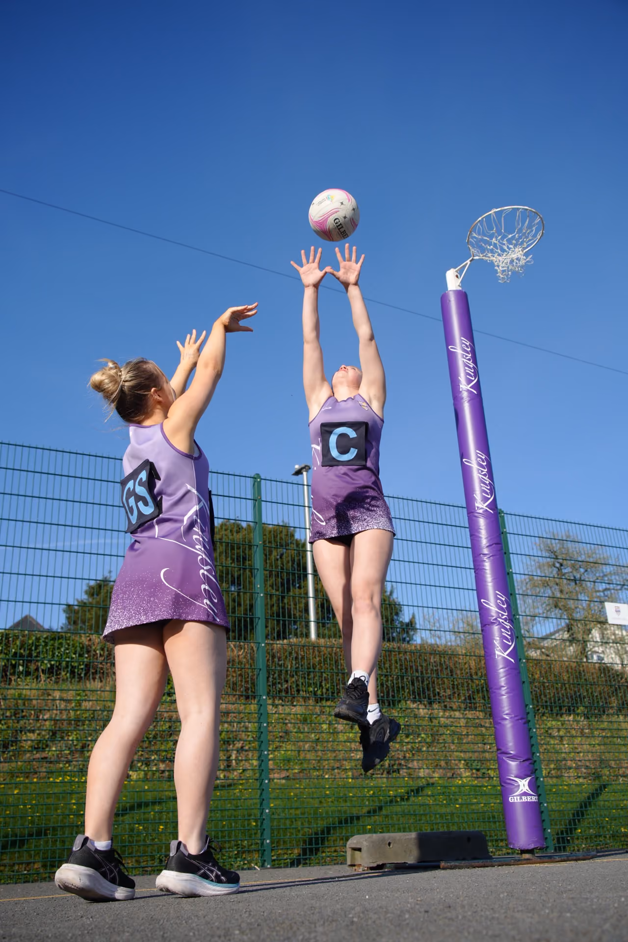 Two female netball players in purple uniforms playing outdoors under a clear blue sky, one jumping to catch a ball near a goal post.