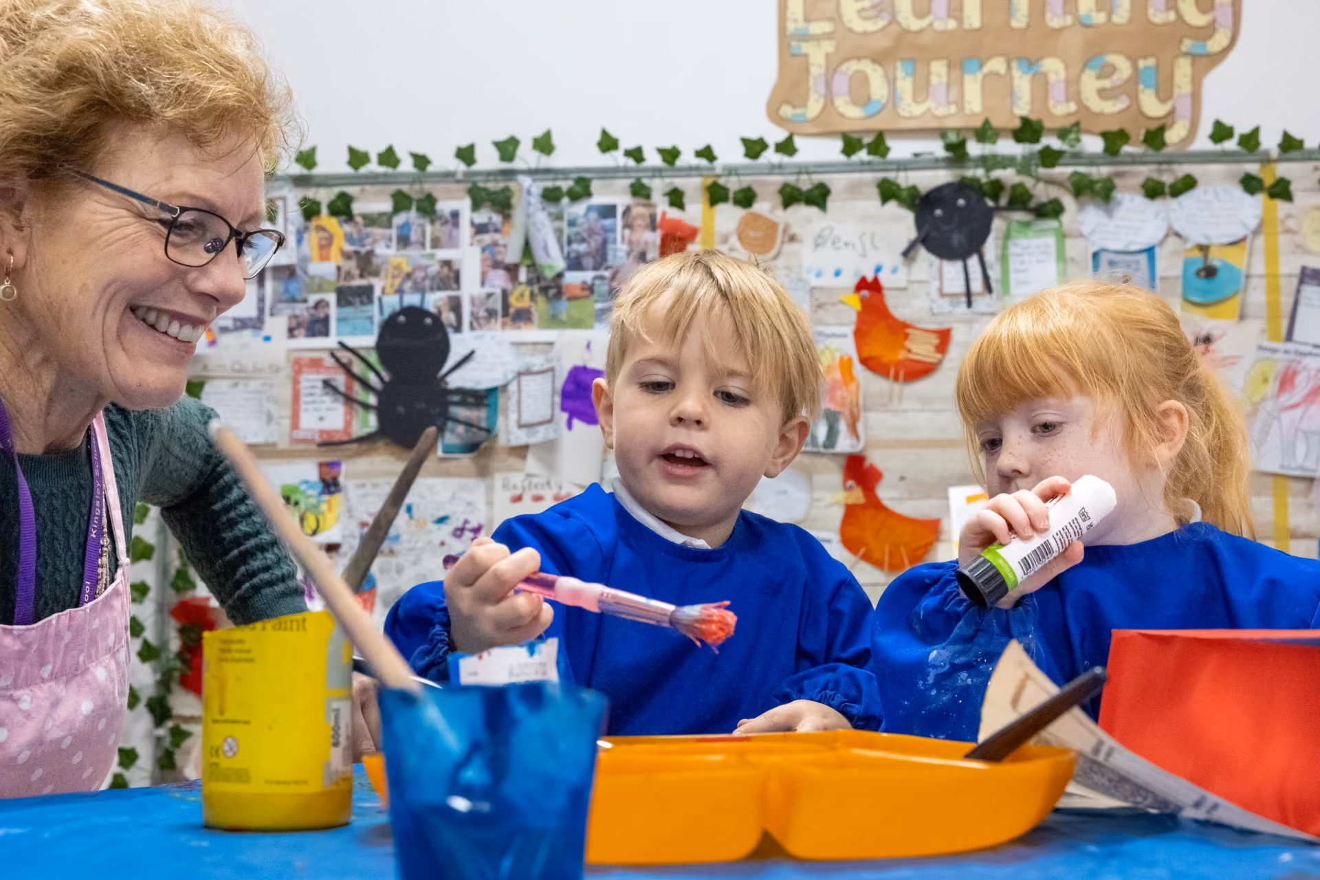 Two young children in blue aprons engage in a painting activity with an older woman smiling beside them in a classroom decorated with children's artwork and a sign reading 'Learning Journey'.