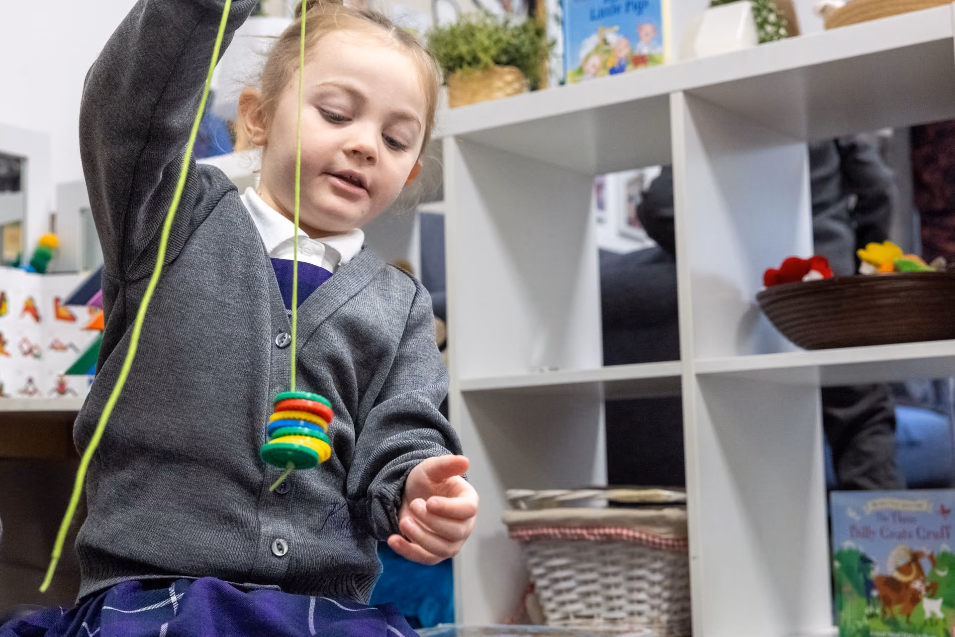 A young girl in a grey cardigan and school uniform playing with a string toy with colorful rings in a classroom or nursery setting.