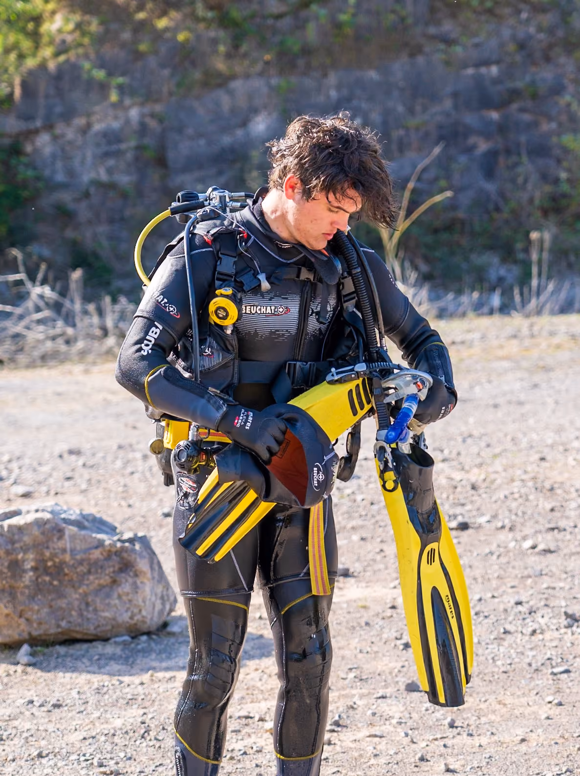 Man wearing a black wetsuit with yellow accents adjusting his yellow and black scuba diving fins outdoors on a rocky surface with greenery in the background.