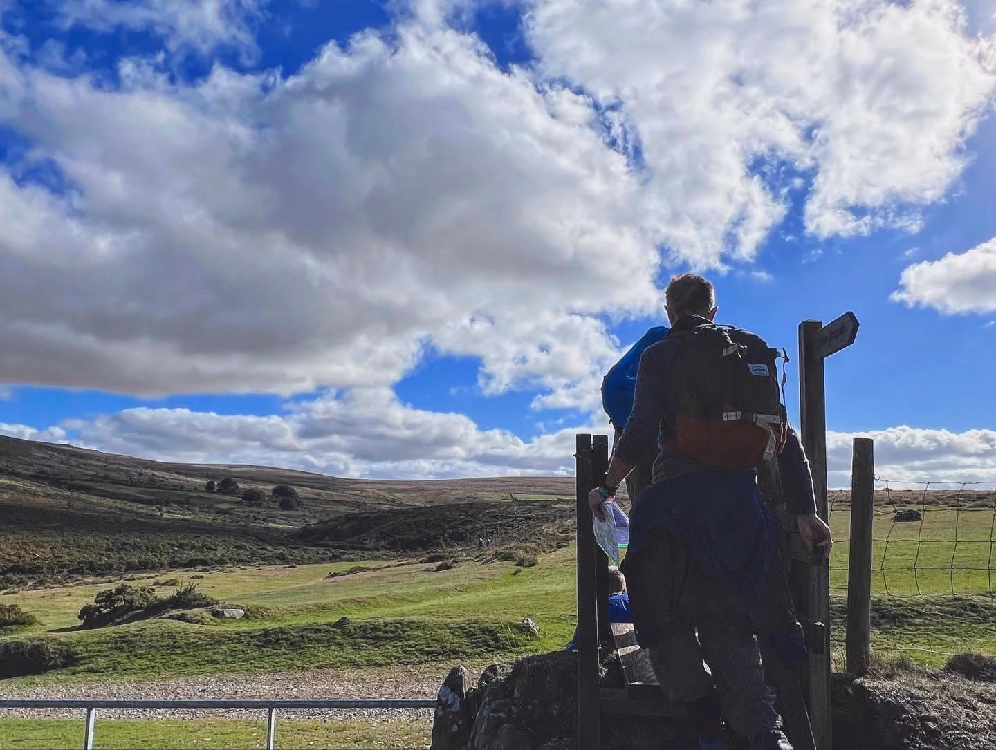 Two hikers climbing a wooden stile over a wire fence in a grassy, hilly landscape under a blue sky with scattered clouds.