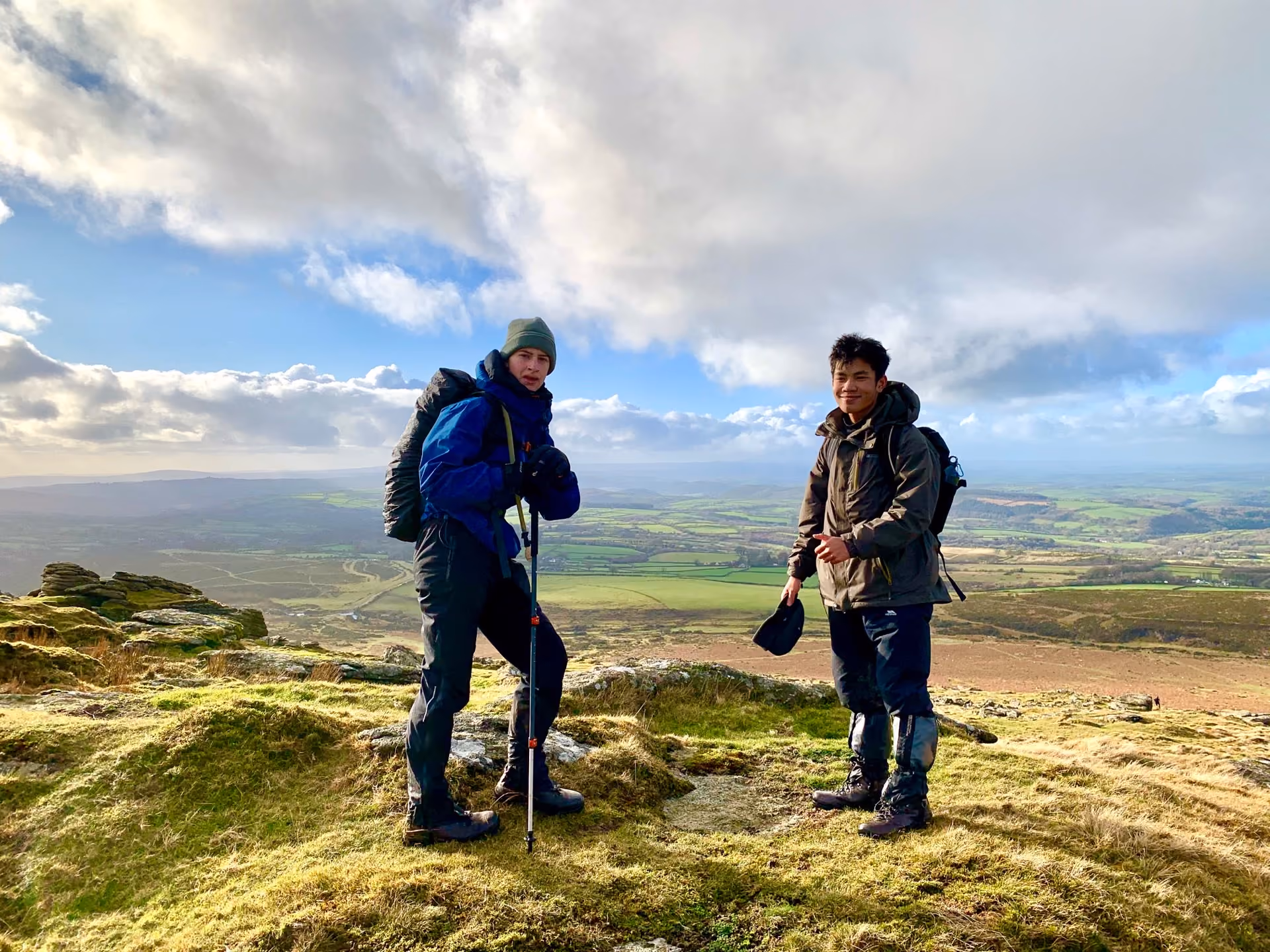 Two hikers standing on a grassy hilltop with hiking gear, overlooking a wide landscape of green fields and hills under a partly cloudy blue sky.