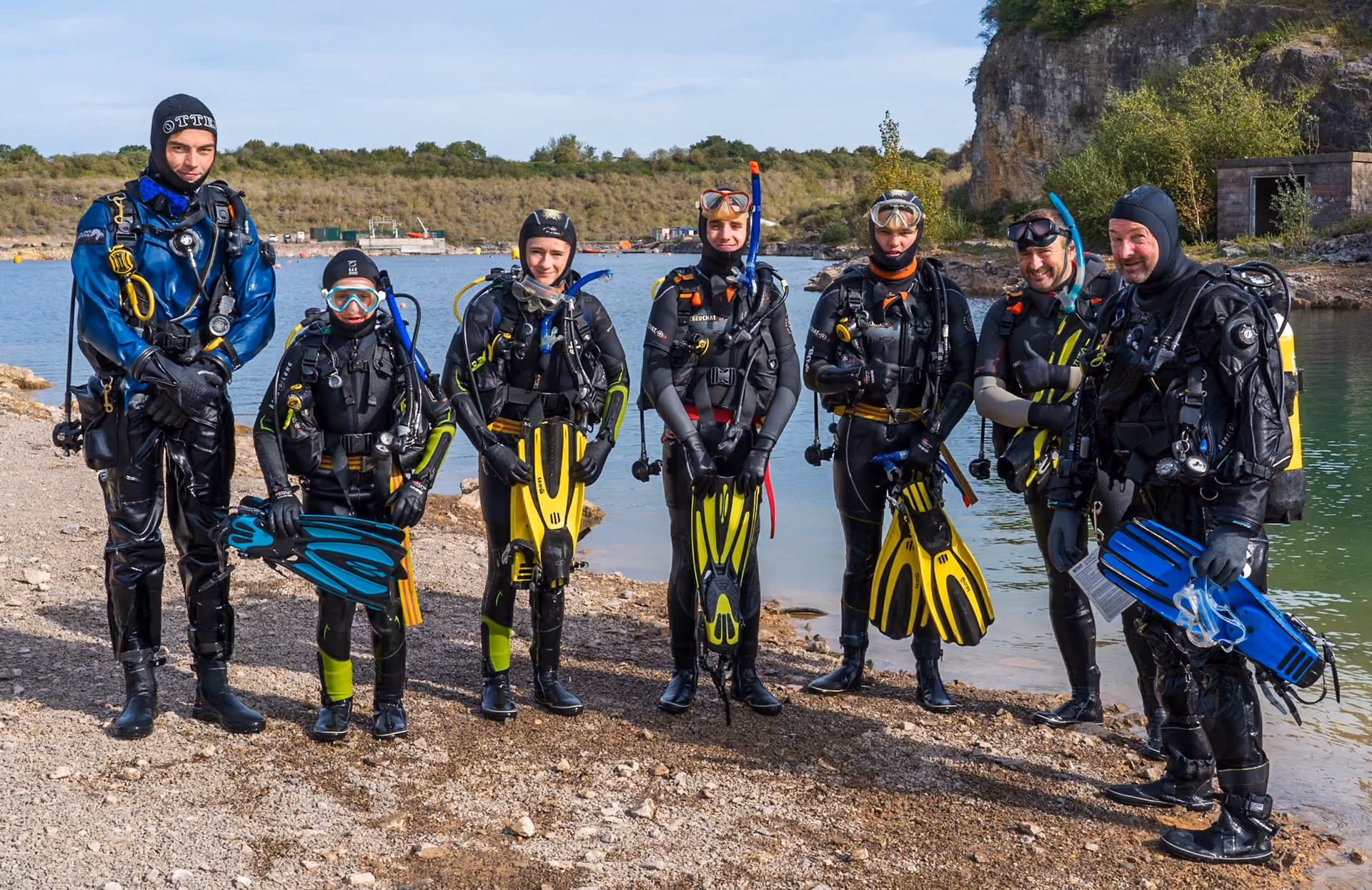 Seven scuba divers standing on a rocky shore near water, wearing diving suits and holding fins, with trees and cliffs in the background.