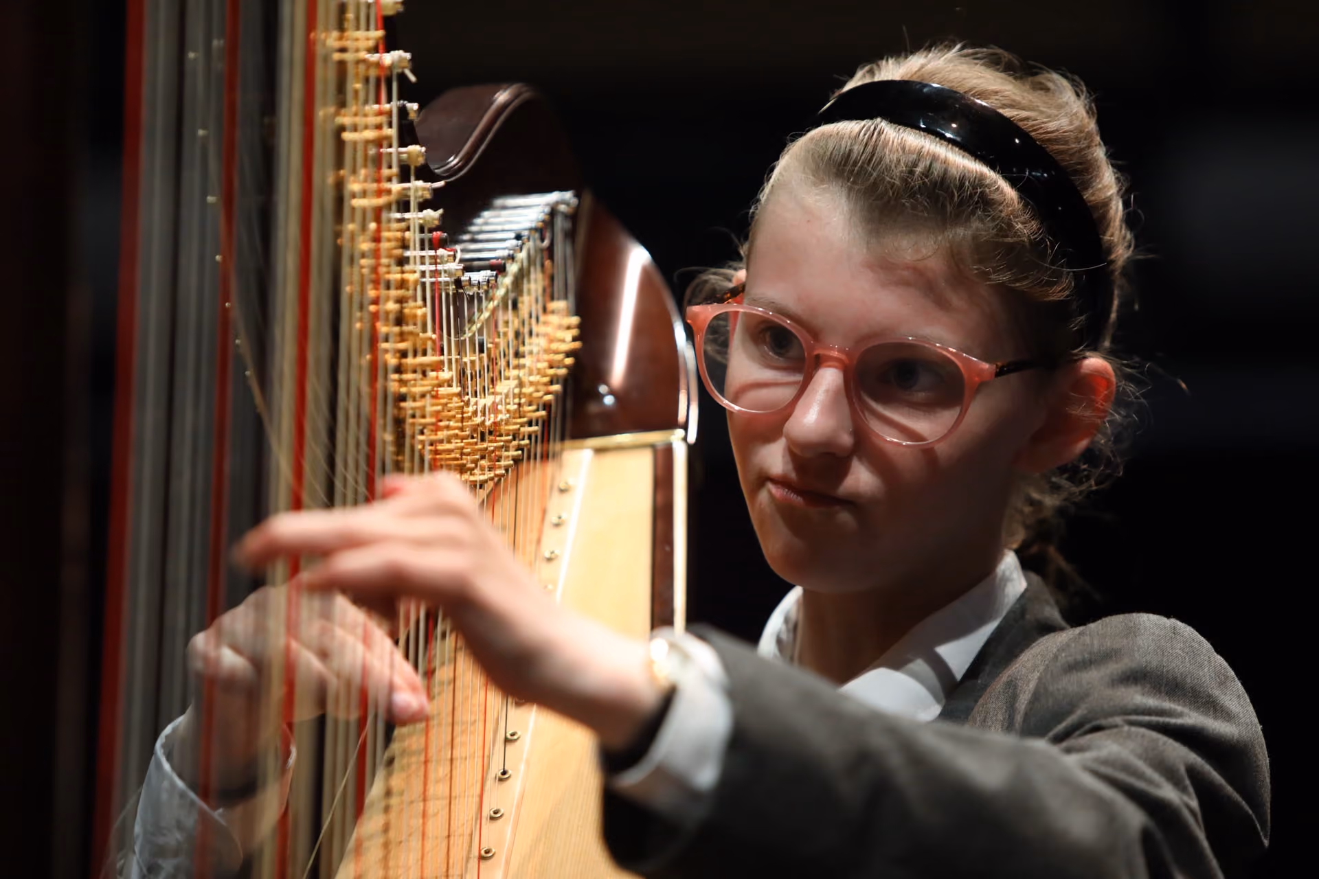 Young woman with red glasses and a black headband focused on playing a large wooden harp with both hands.