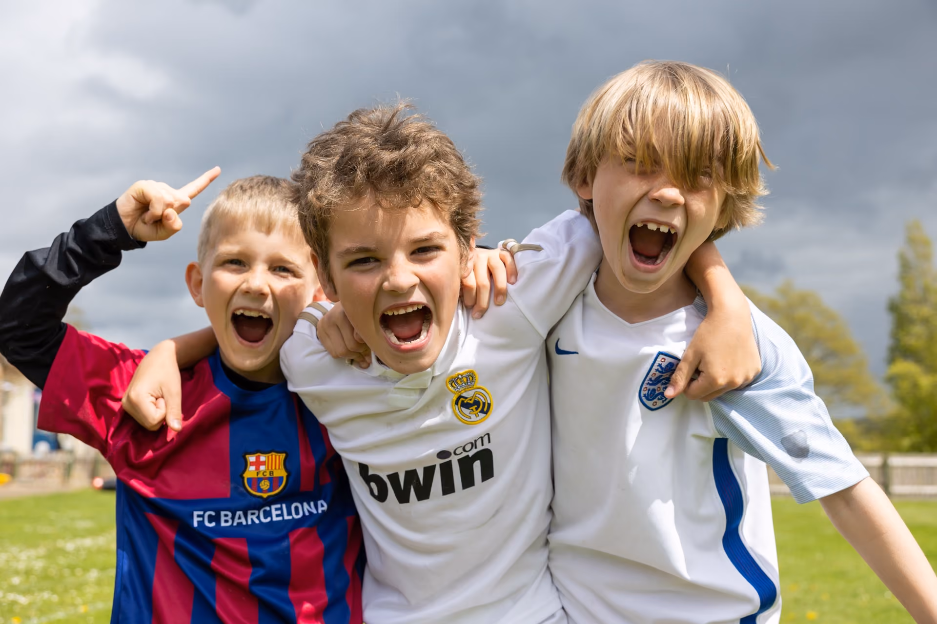 Three boys outdoors hugging and cheering, wearing football shirts of FC Barcelona, Real Madrid, and England, with a cloudy sky in the background.