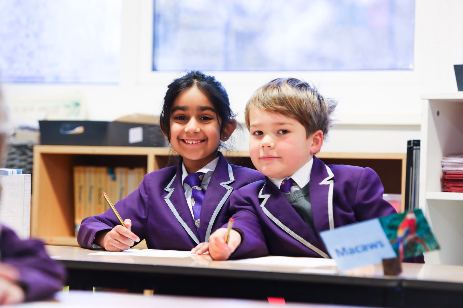 Two primary school children in purple blazers with white trim sitting at a classroom desk, smiling and holding pencils.