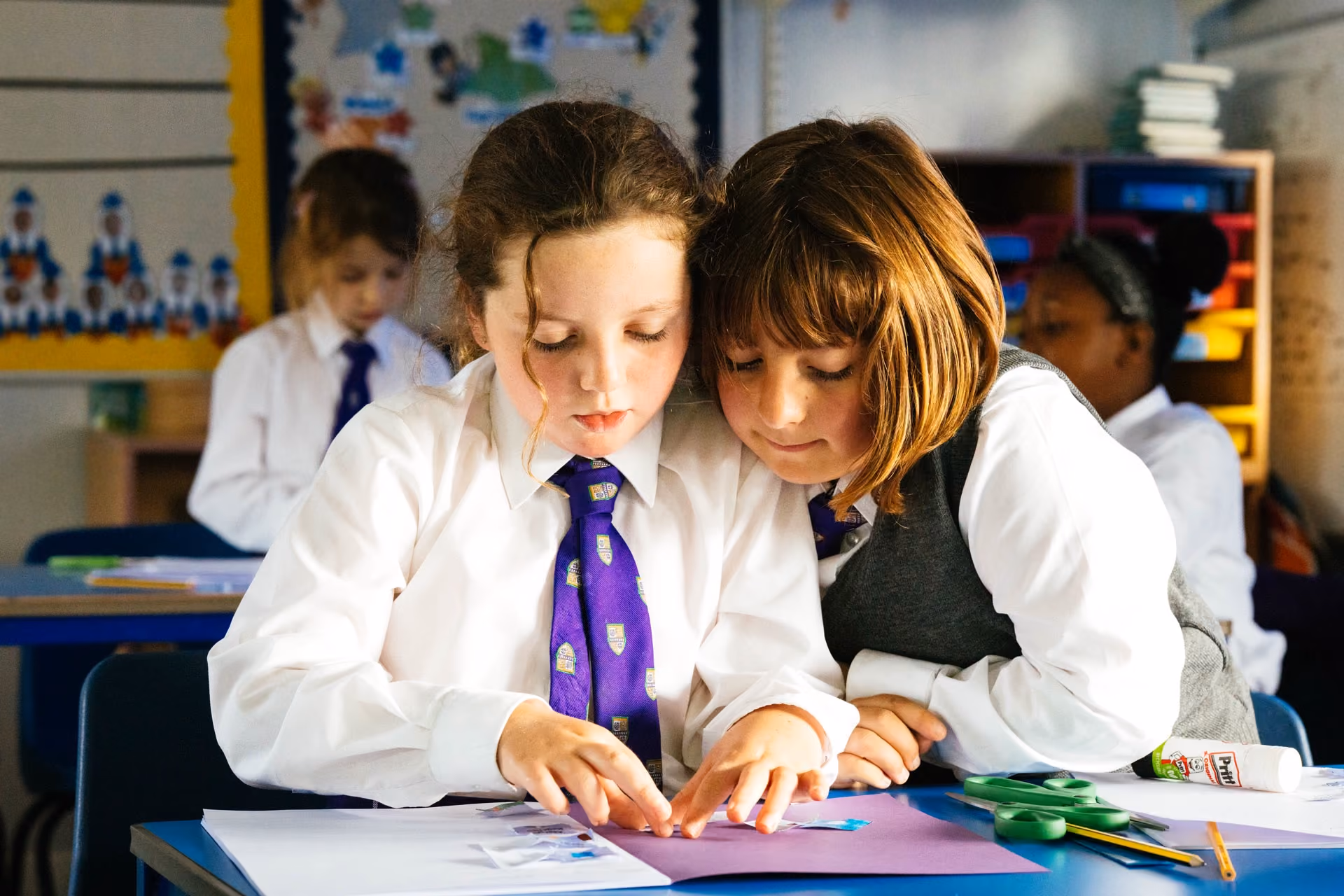 Two schoolgirls in uniform working together on a craft project at a desk, one pointing at paper with glue sticks and scissors nearby, with other students blurred in the background.