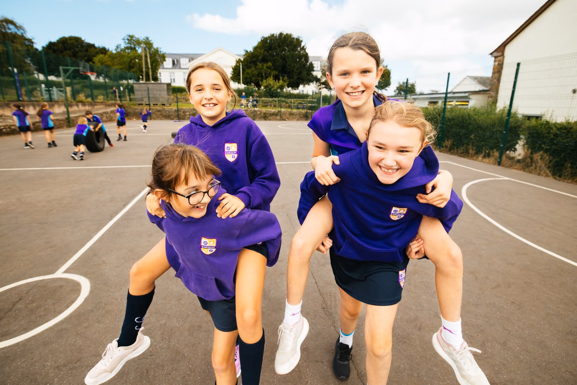 Four schoolgirls outdoors on a sports court, two girls giving piggyback rides to the other two, all smiling and wearing purple school jumpers with badges and dark skirts, with other children playing in the background.