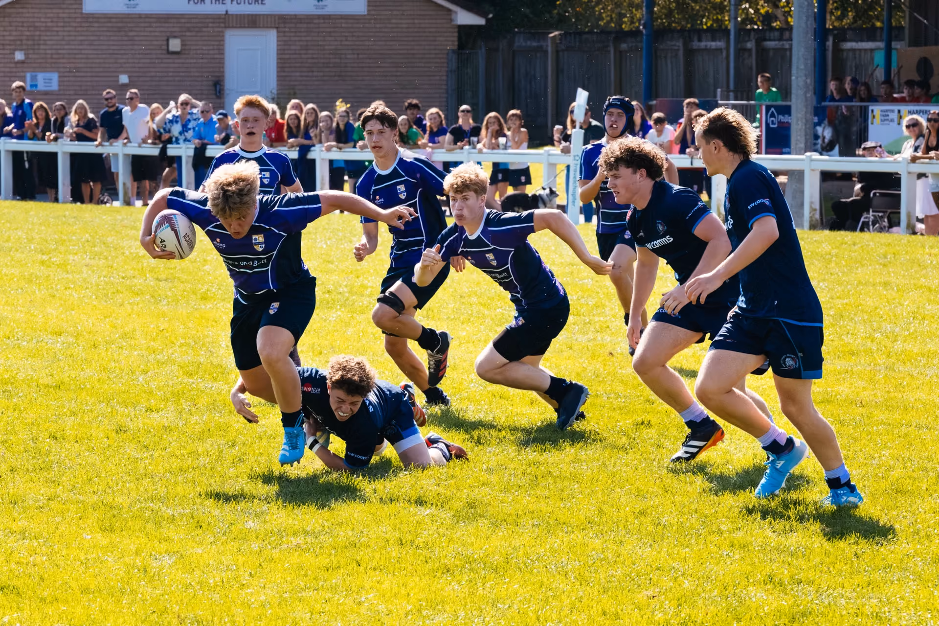 Youth rugby players in blue uniforms playing on a sunny field, with one player running while holding the ball and another attempting a tackle, spectators visible in the background behind a white fence.