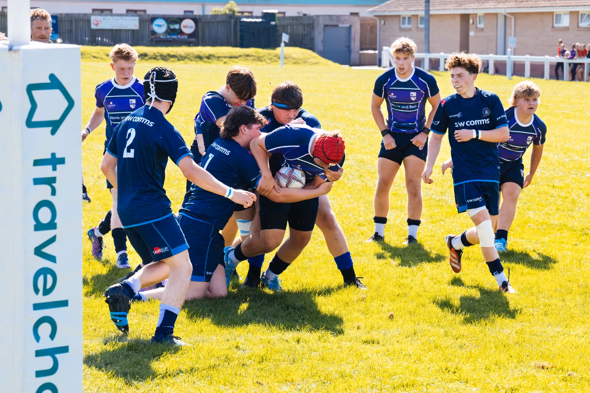 A group of youth rugby players in blue uniforms engaged in a match on a sunny field, with one player holding the ball and others attempting to tackle or support him near a goal post featuring a travelcho logo.
