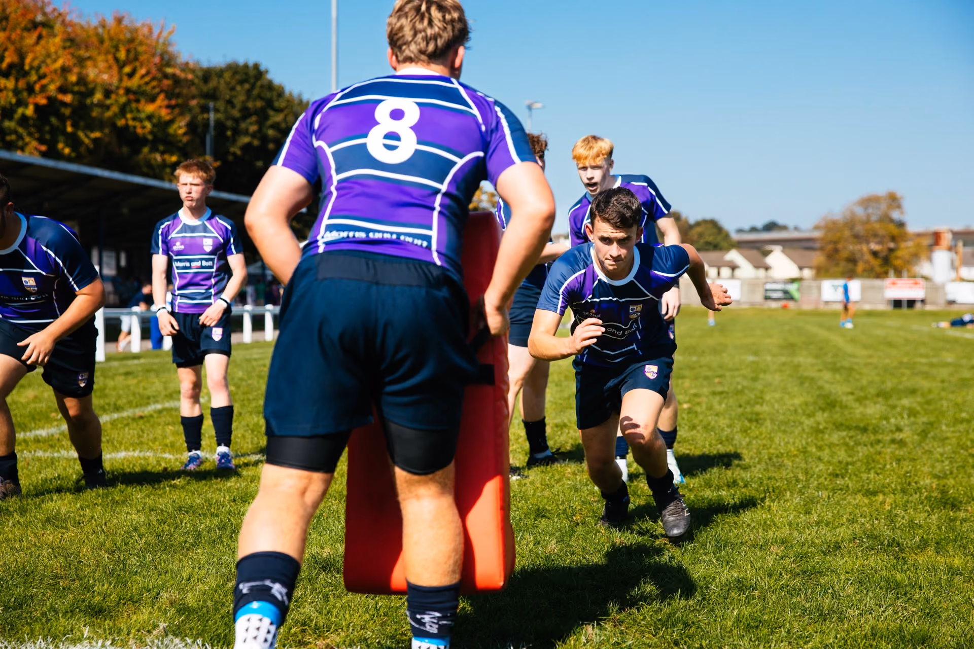 Young rugby players in purple and blue jerseys practicing on a grassy field under clear blue sky, one player running towards a teammate holding a red tackling pad.