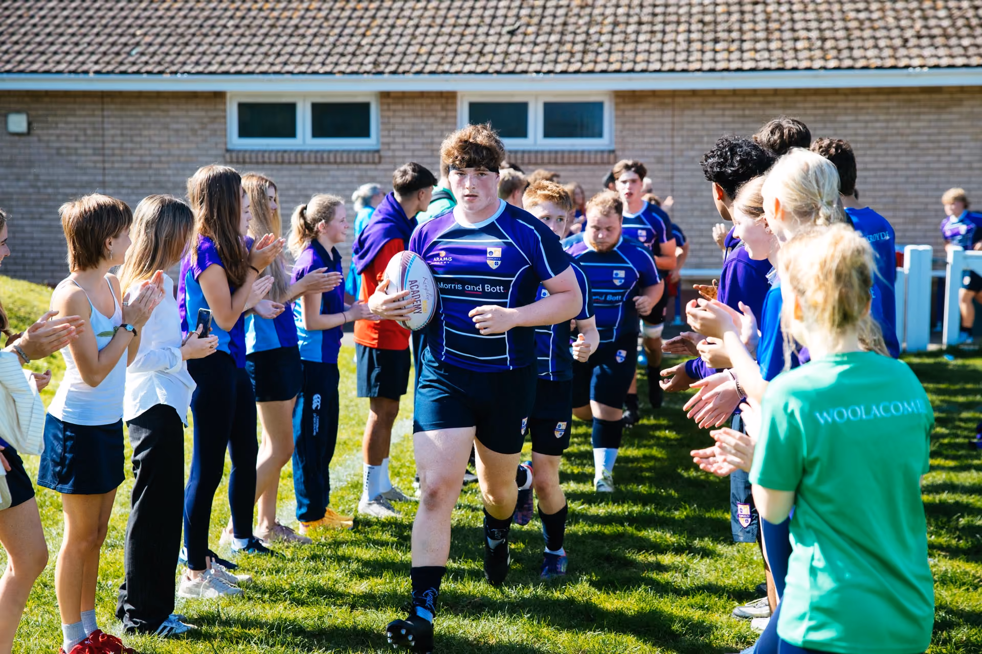 Young rugby players in blue uniforms run on a grassy field through a corridor formed by applauding students on either side, with a brick building in the background.