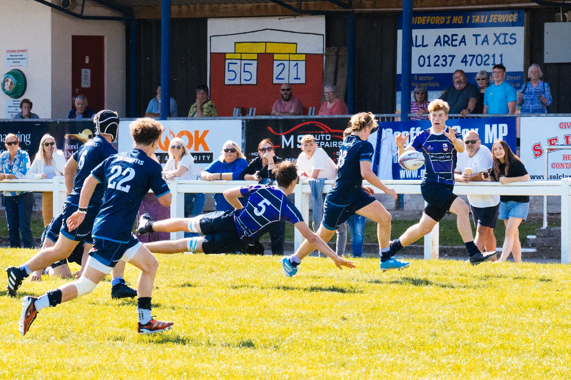 A rugby match in progress with players in blue and black kits actively running and diving on a sunny grassy field, with spectators watching behind a white barrier and a scoreboard showing 55 to 21 in the background.
