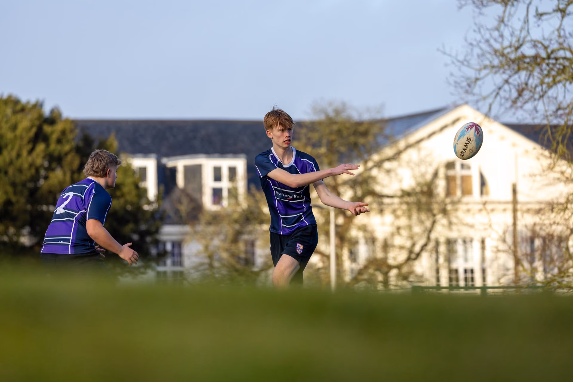 Two young rugby players in purple and blue uniforms on a grassy field with a building and trees in the background, one player passing a rugby ball to the other.