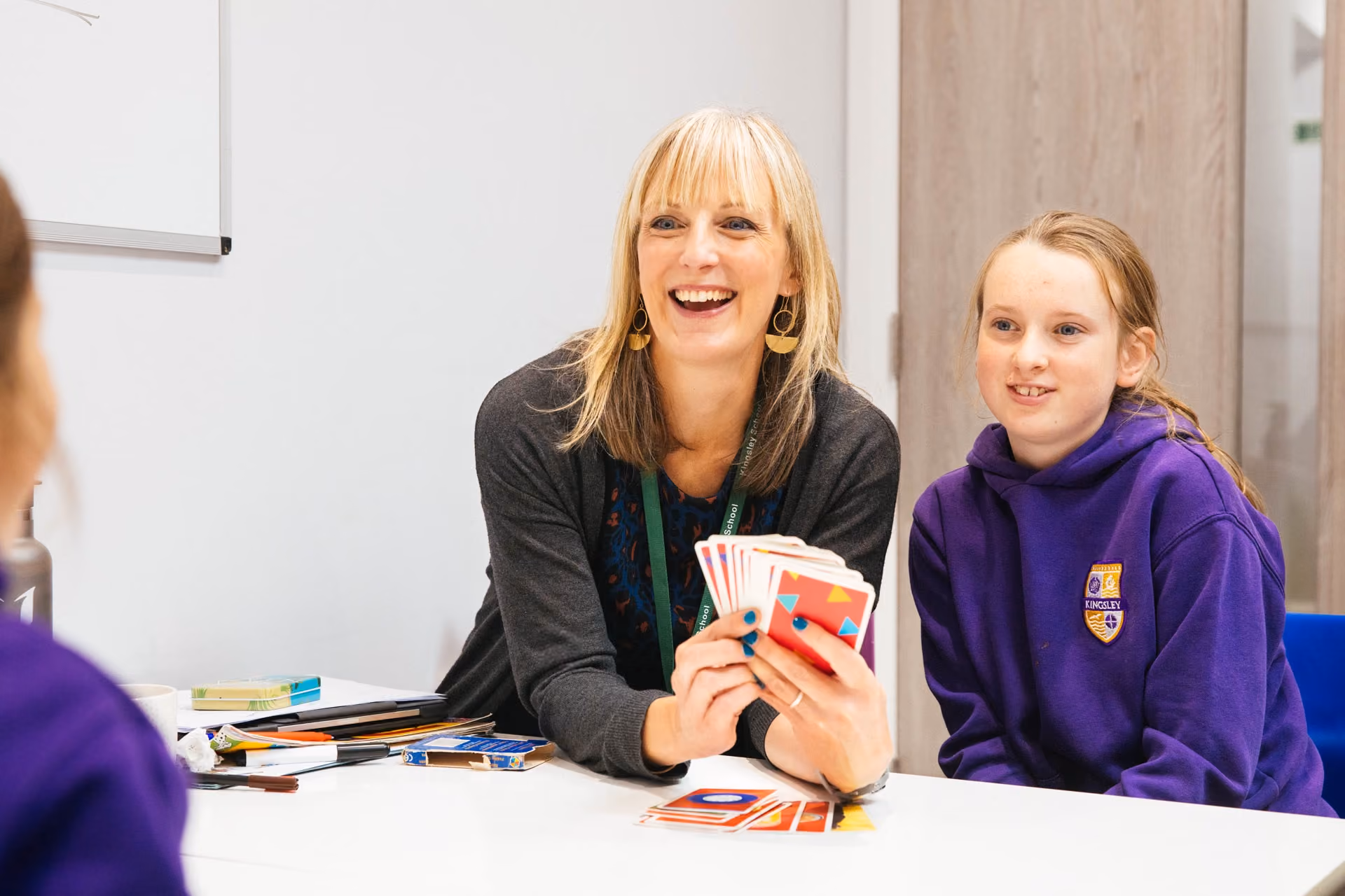 Smiling woman holding colorful cards in her hands sitting at a table with a girl in a purple school uniform hoodie, both engaged and happy during an indoor activity.