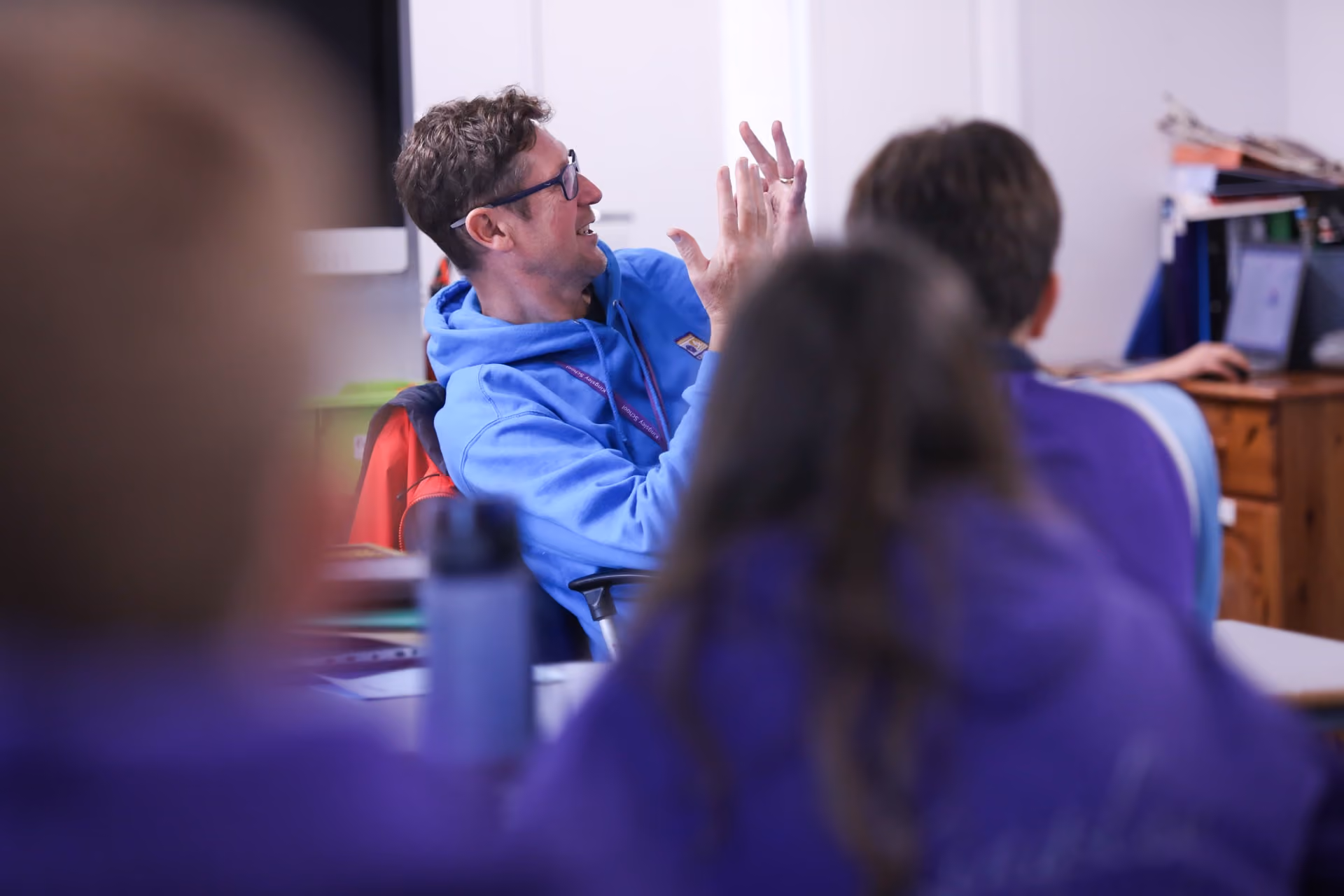 A man wearing glasses and a blue hoodie is animatedly gesturing and speaking to a group of people seated in a classroom setting.