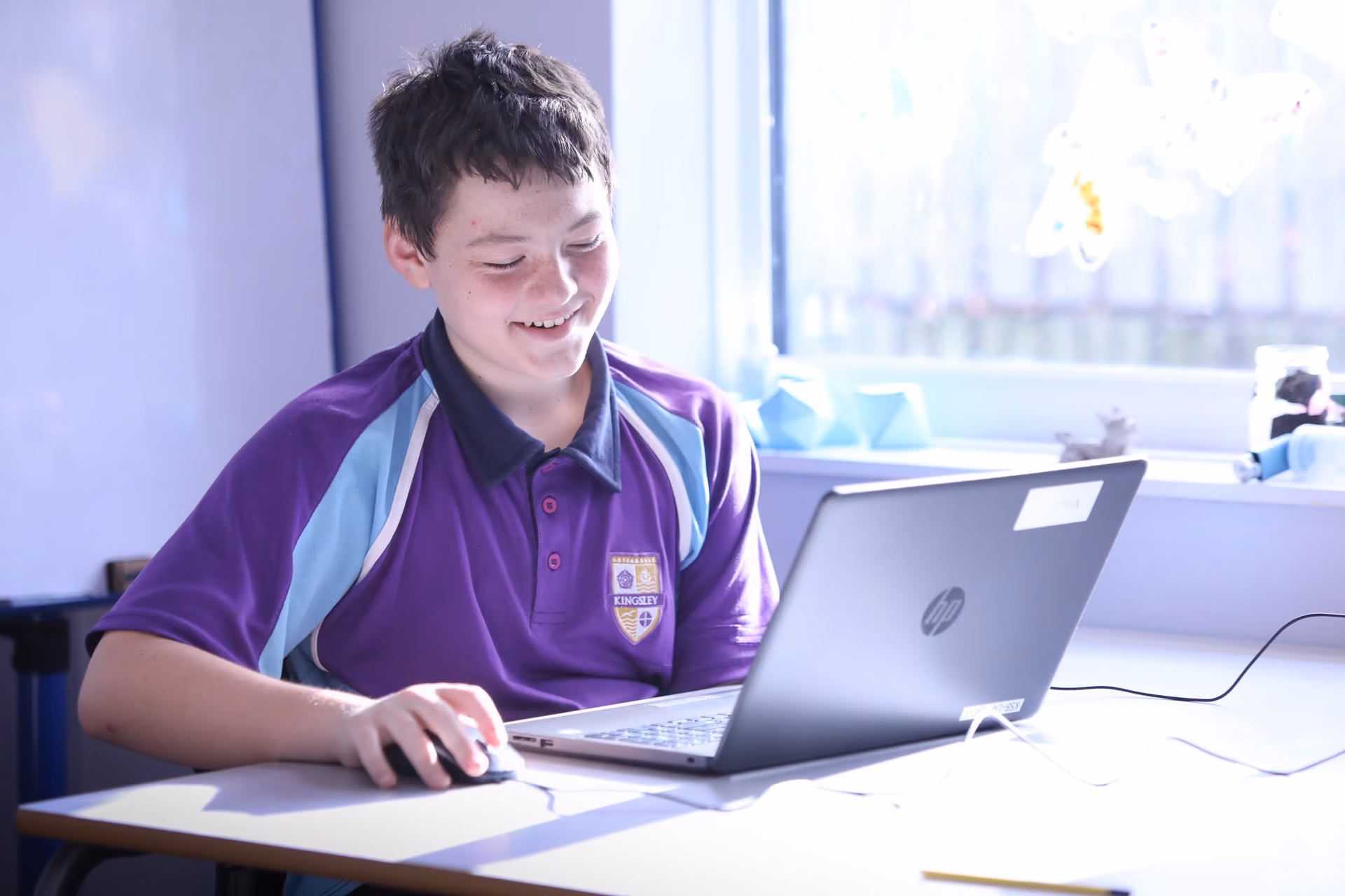 A smiling boy wearing a purple and light blue school polo shirt with a badge, using a laptop at a desk by a window in a bright room.