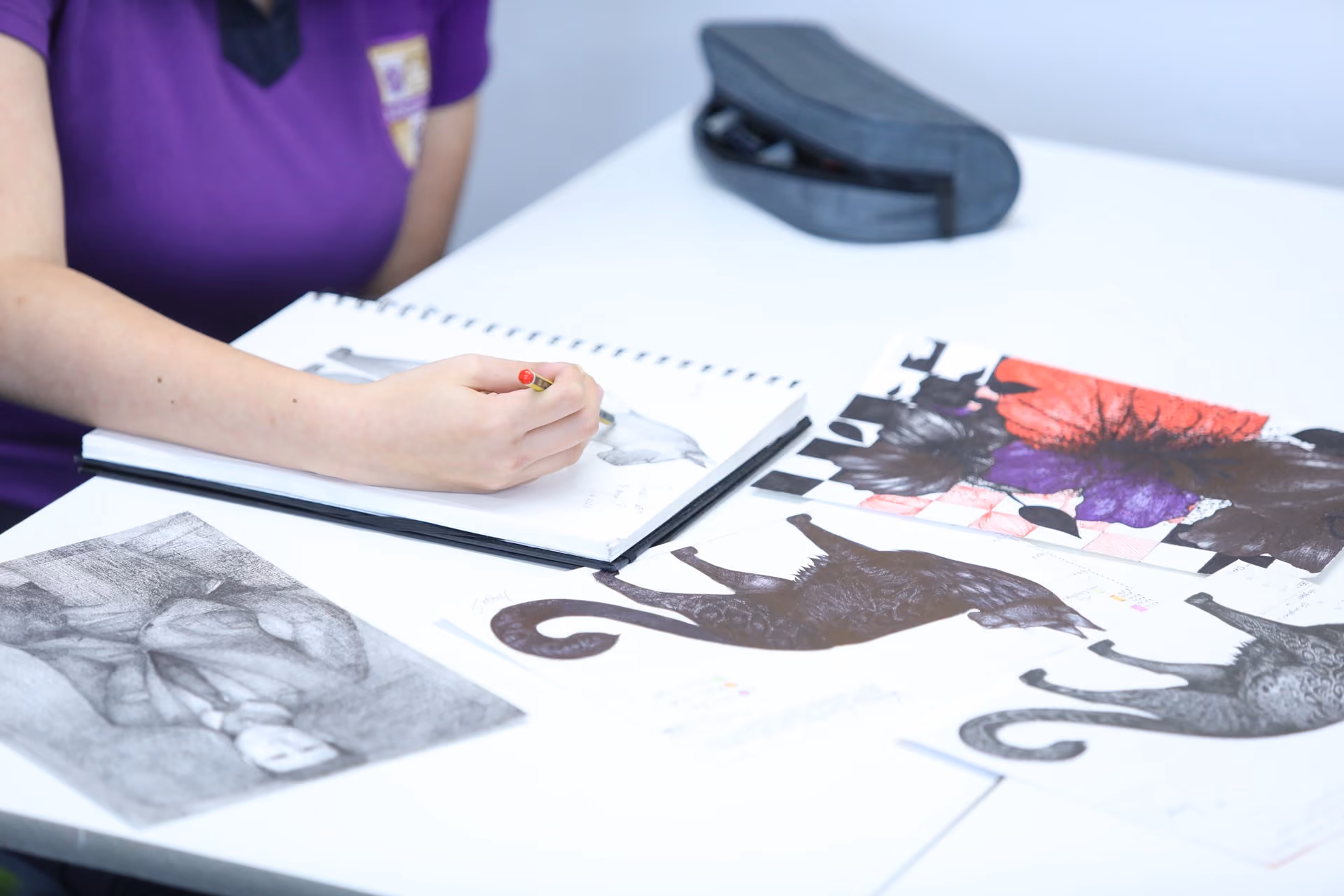 Person in a purple top sketching on a pad at a white table, surrounded by various drawings of cats, including black, white, and multicoloured designs.