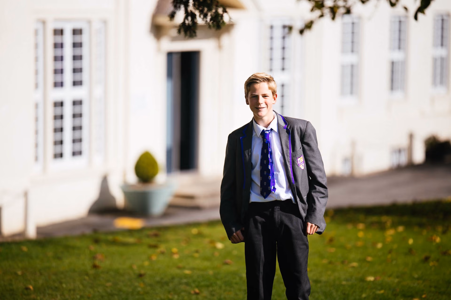 Teenage boy in a school uniform with a grey blazer and purple striped tie standing on grass outside a white building with windows.