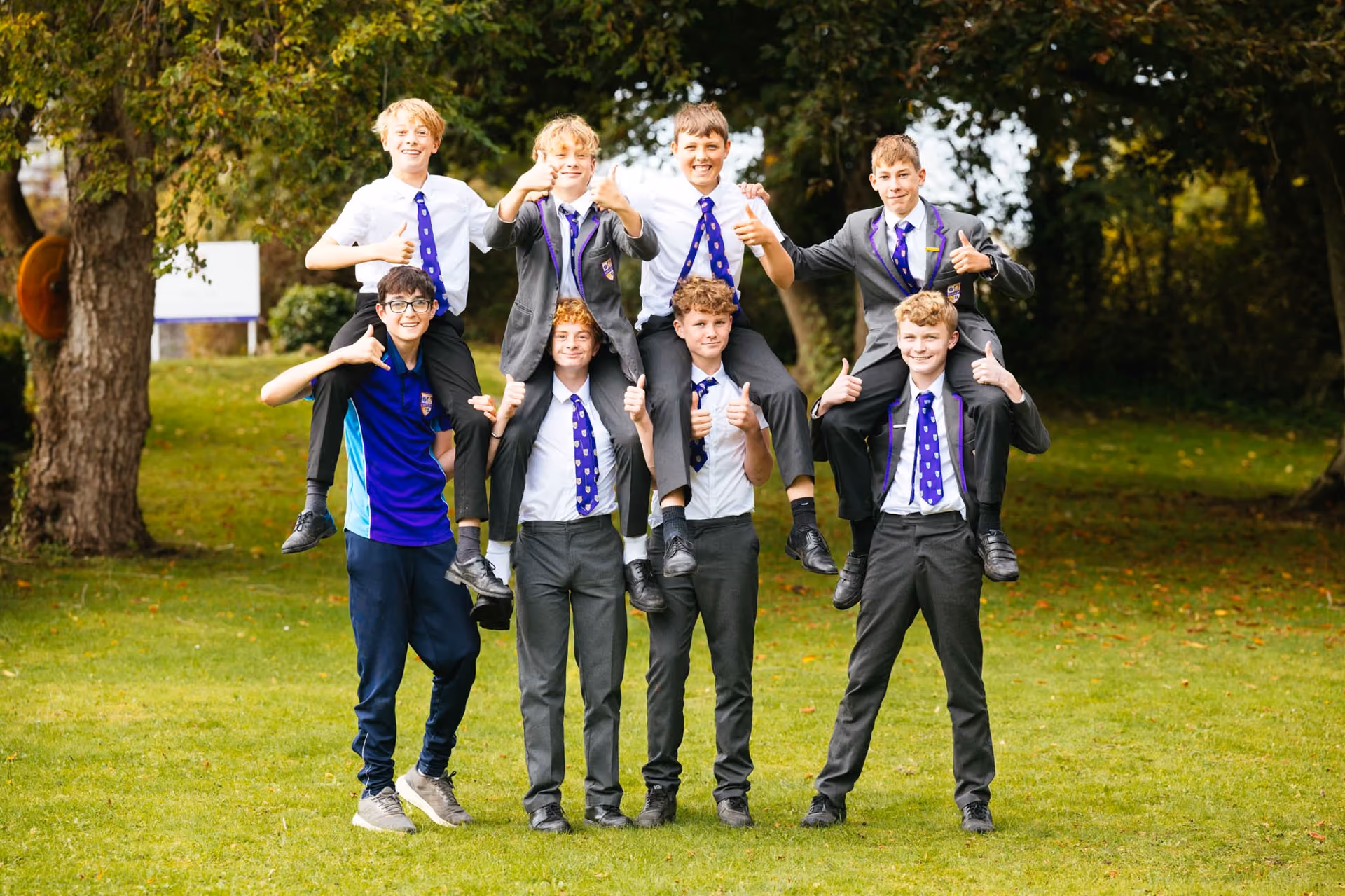 A group of eight schoolboys in uniform standing on grass with trees in the background; four boys are carrying four others on their shoulders, all smiling and giving thumbs-up.