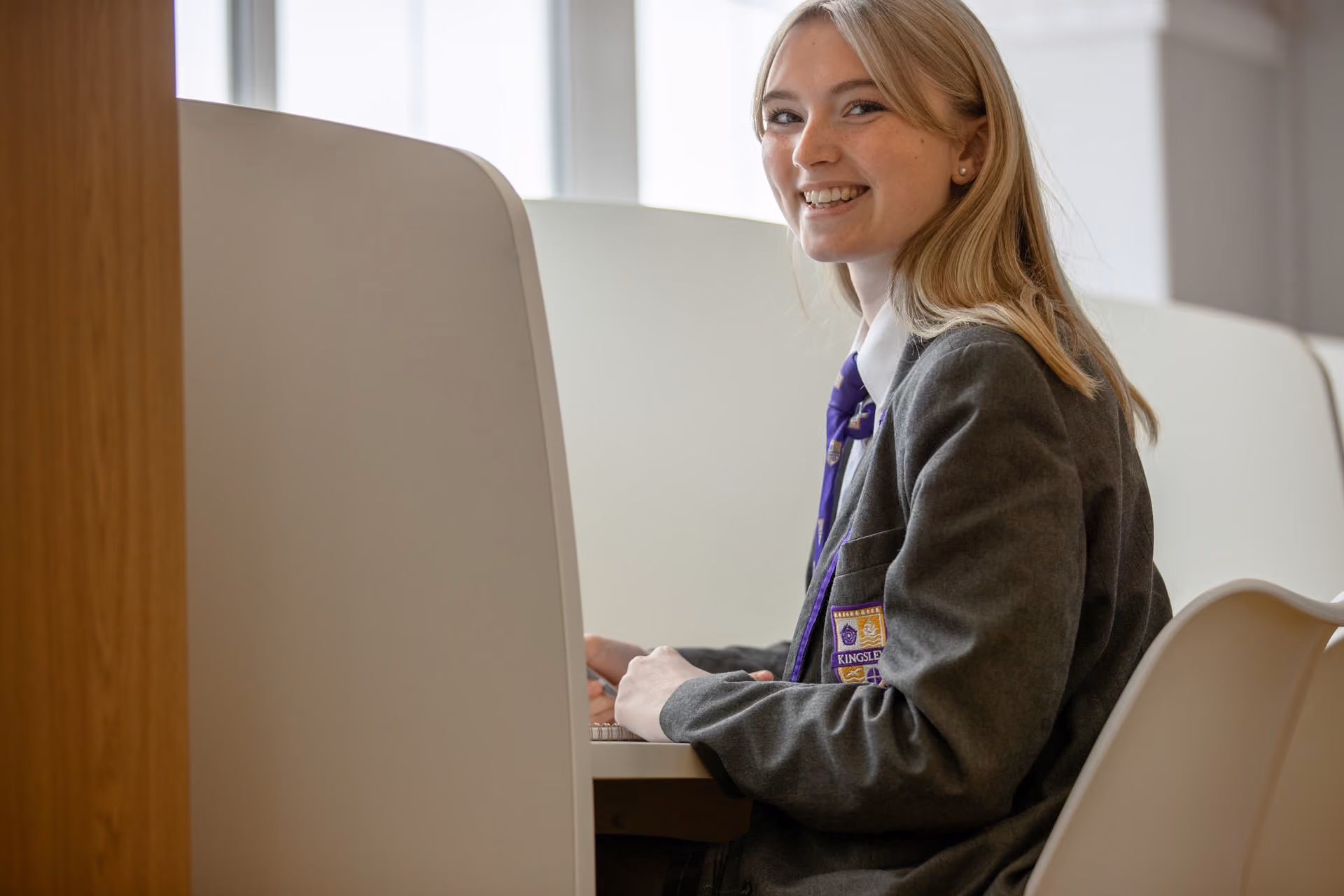 Smiling young woman in a grey school blazer with a purple and gold emblem and tie, sitting at a desk in a bright room with large windows.