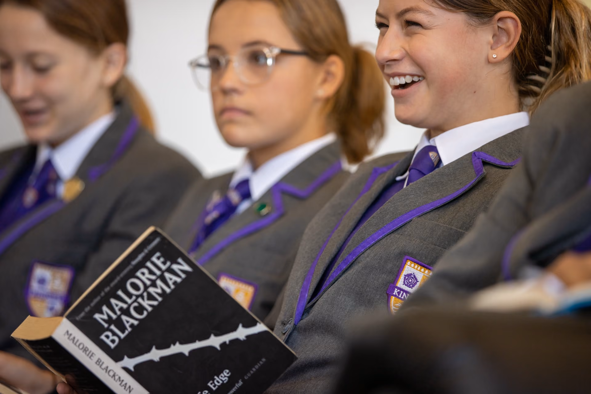 Three schoolgirls in grey uniforms with purple trim sitting together, one smiling and holding an open book titled 'Malorie Blackman, The Edge'.