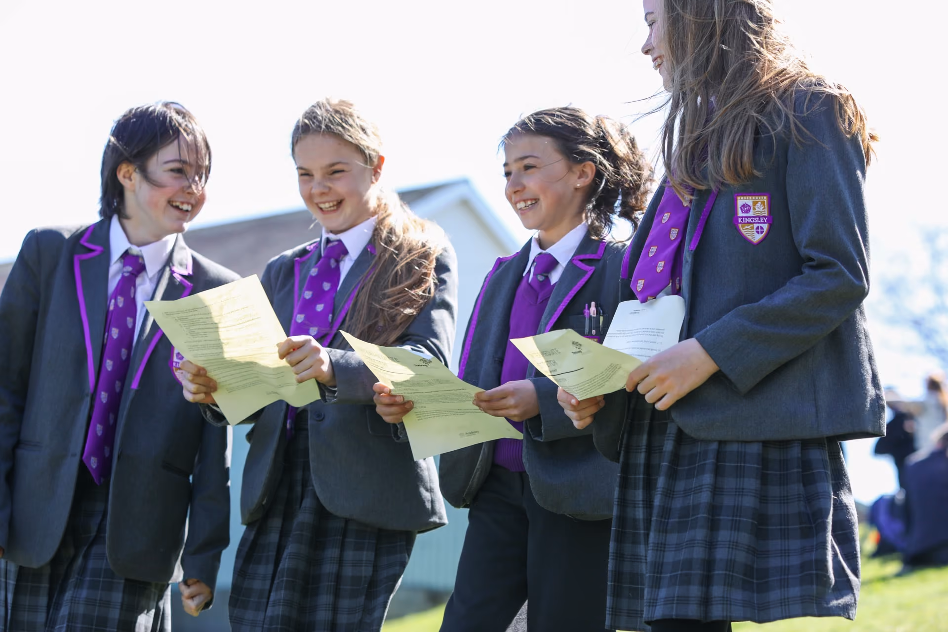 Four schoolgirls in grey blazers with purple ties and plaid skirts standing outside on grass, smiling and holding papers.