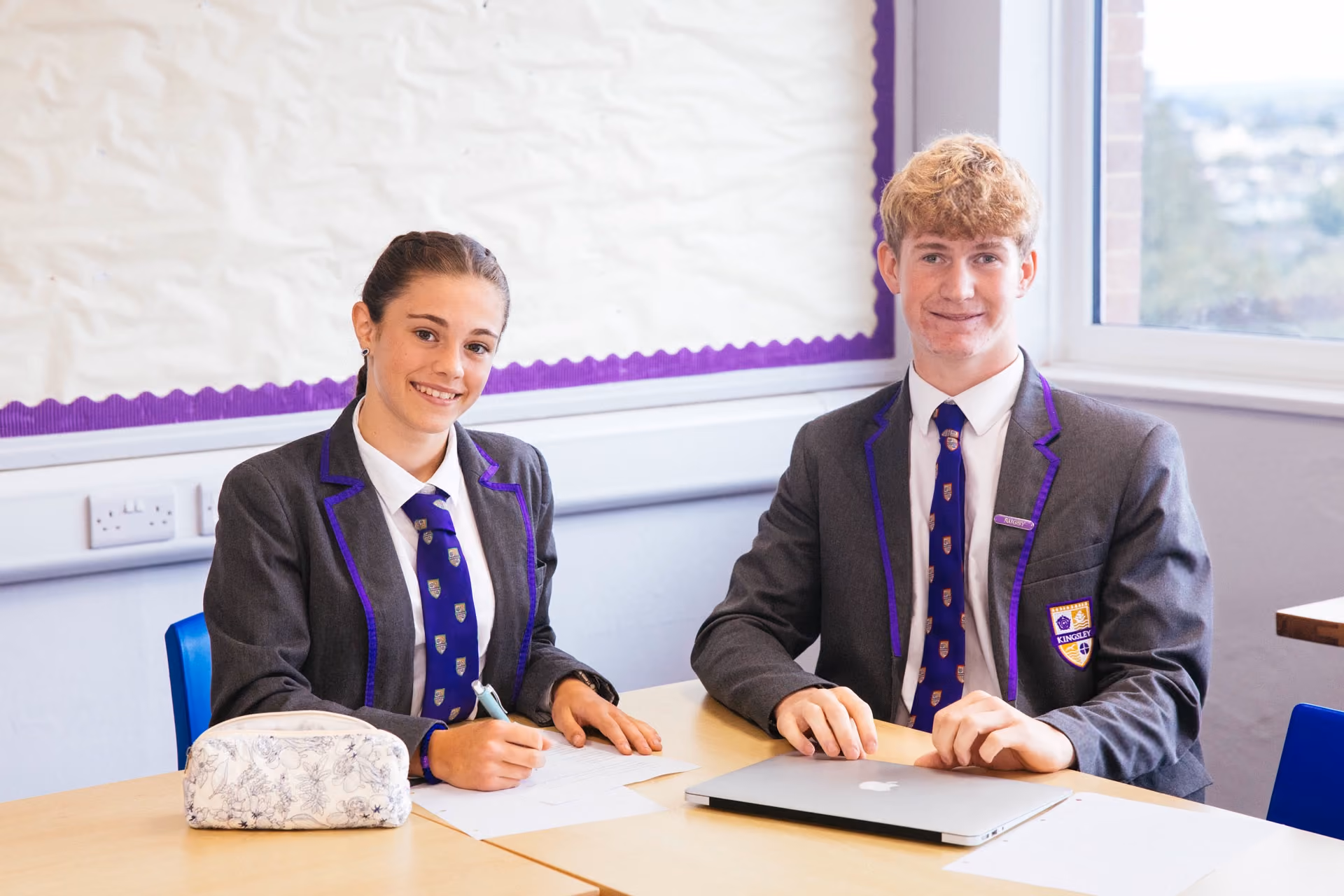 Two students in grey school uniforms with purple trim sit at a desk in a classroom near a window; the girl is writing on paper while the boy sits with a closed laptop.