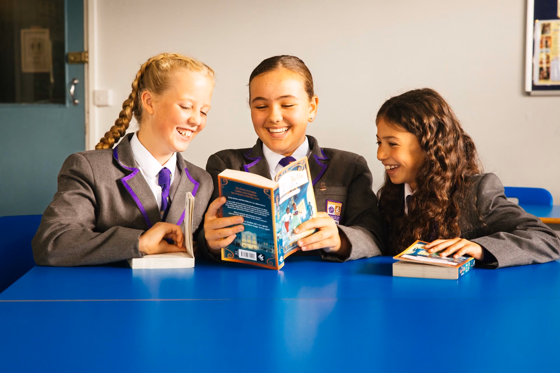 Three teenage girls in school uniforms sitting at a blue table, smiling and reading books together in a classroom.