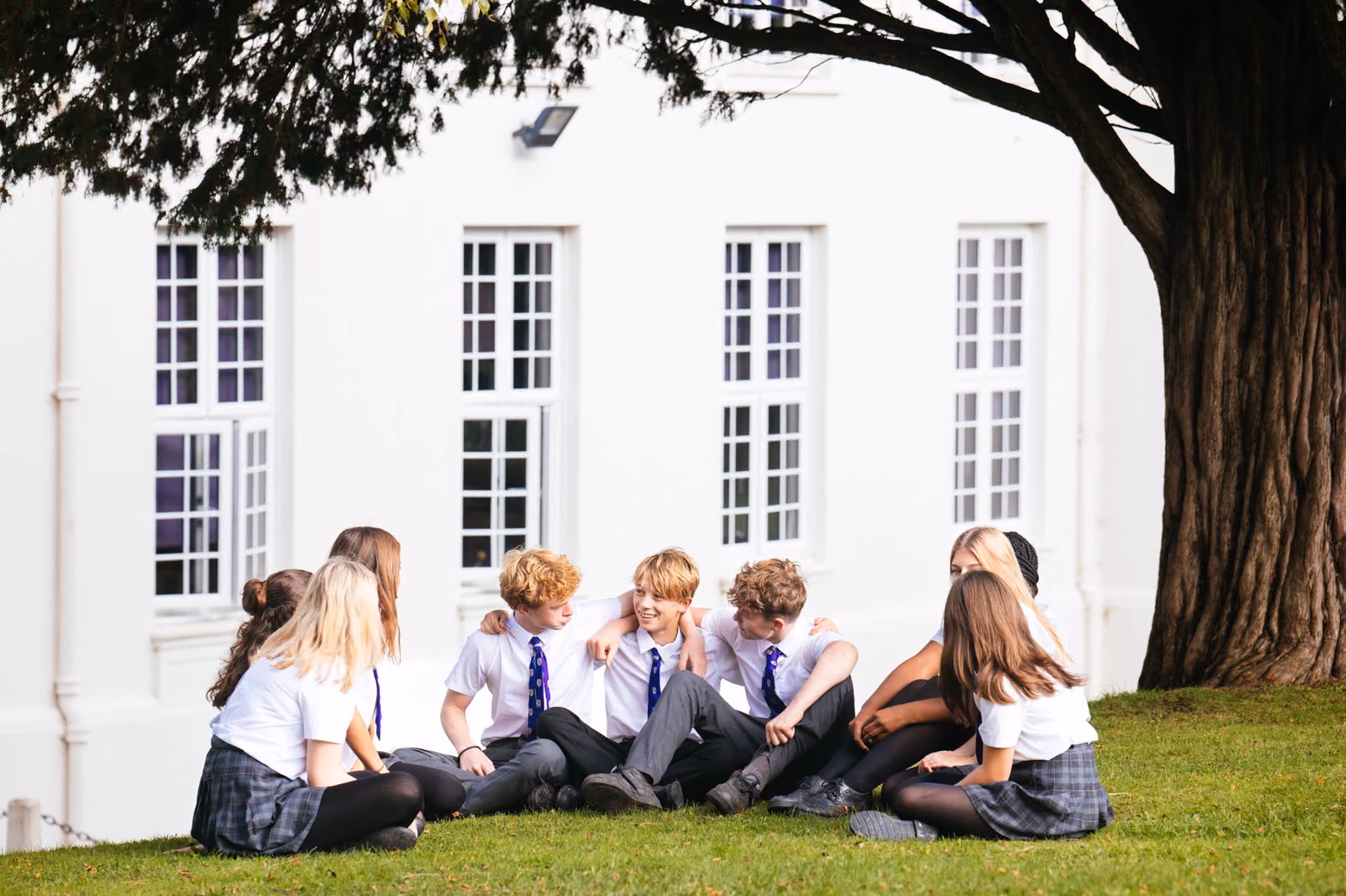 A group of nine schoolchildren dressed in white shirts, ties, and grey checkered skirts or trousers sitting on grass beneath a large tree outside a white building with tall windows, engaging in conversation and smiling.
