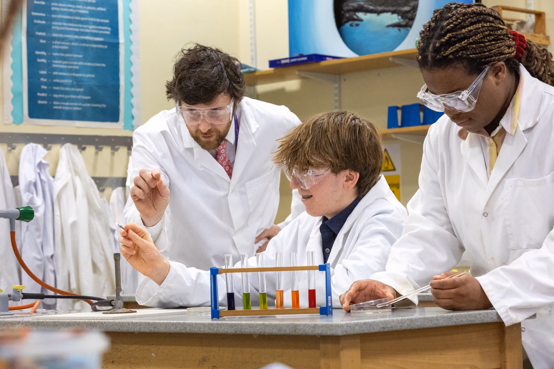 Three people in white lab coats and safety goggles conducting a science experiment with colorful liquids in test tubes on a lab counter, in a classroom setting.