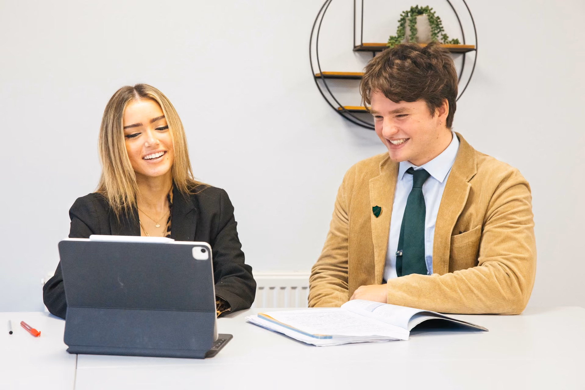 A young woman and a young man sitting side by side at a table, smiling and looking at a tablet device and an open book in front of them, with a circular wall shelf and a small plant in the background.