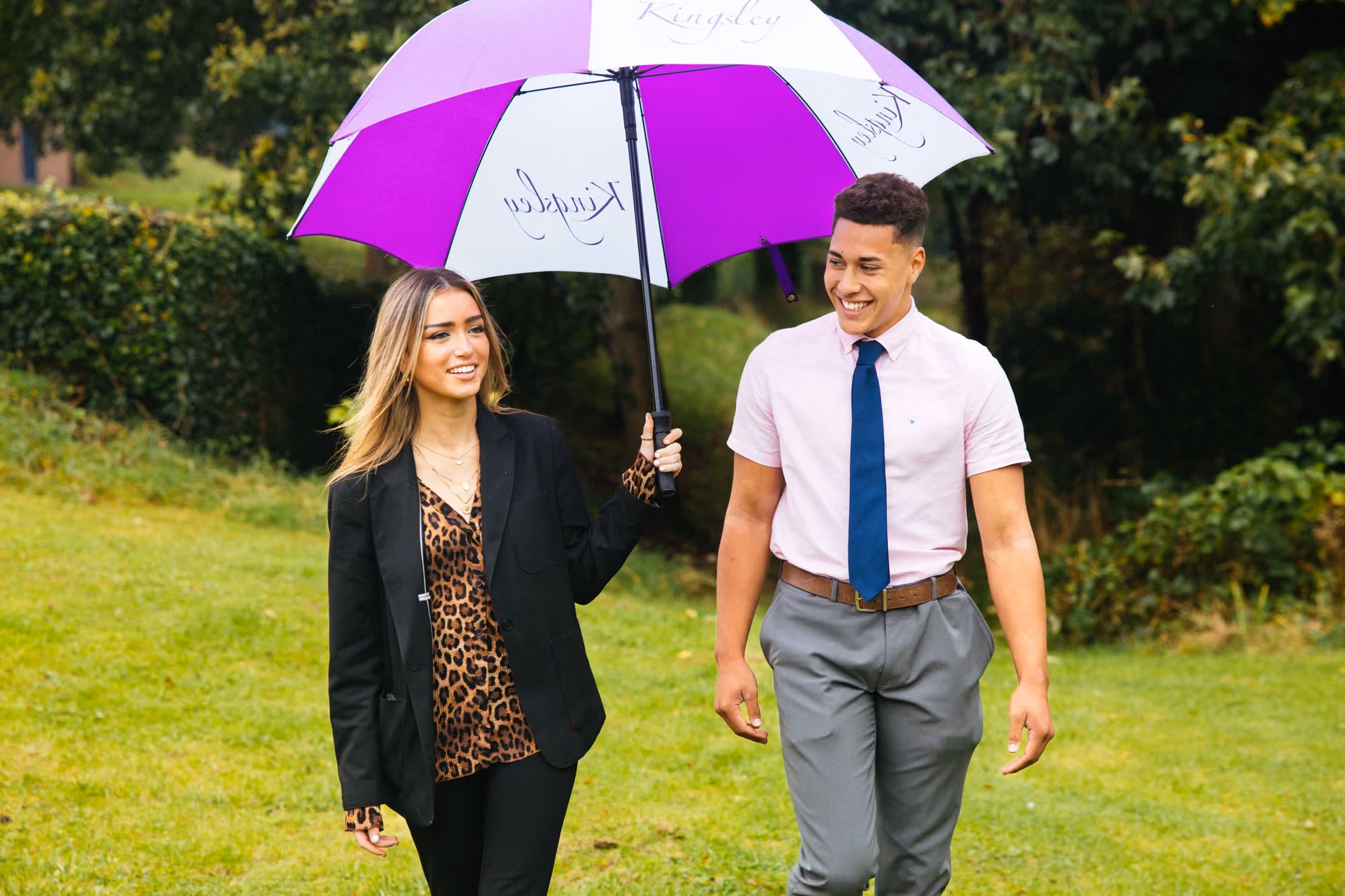A smiling young woman holding a purple and white umbrella walks alongside a young man in grey trousers, pink shirt, and blue tie on a grassy path with trees in the background.
