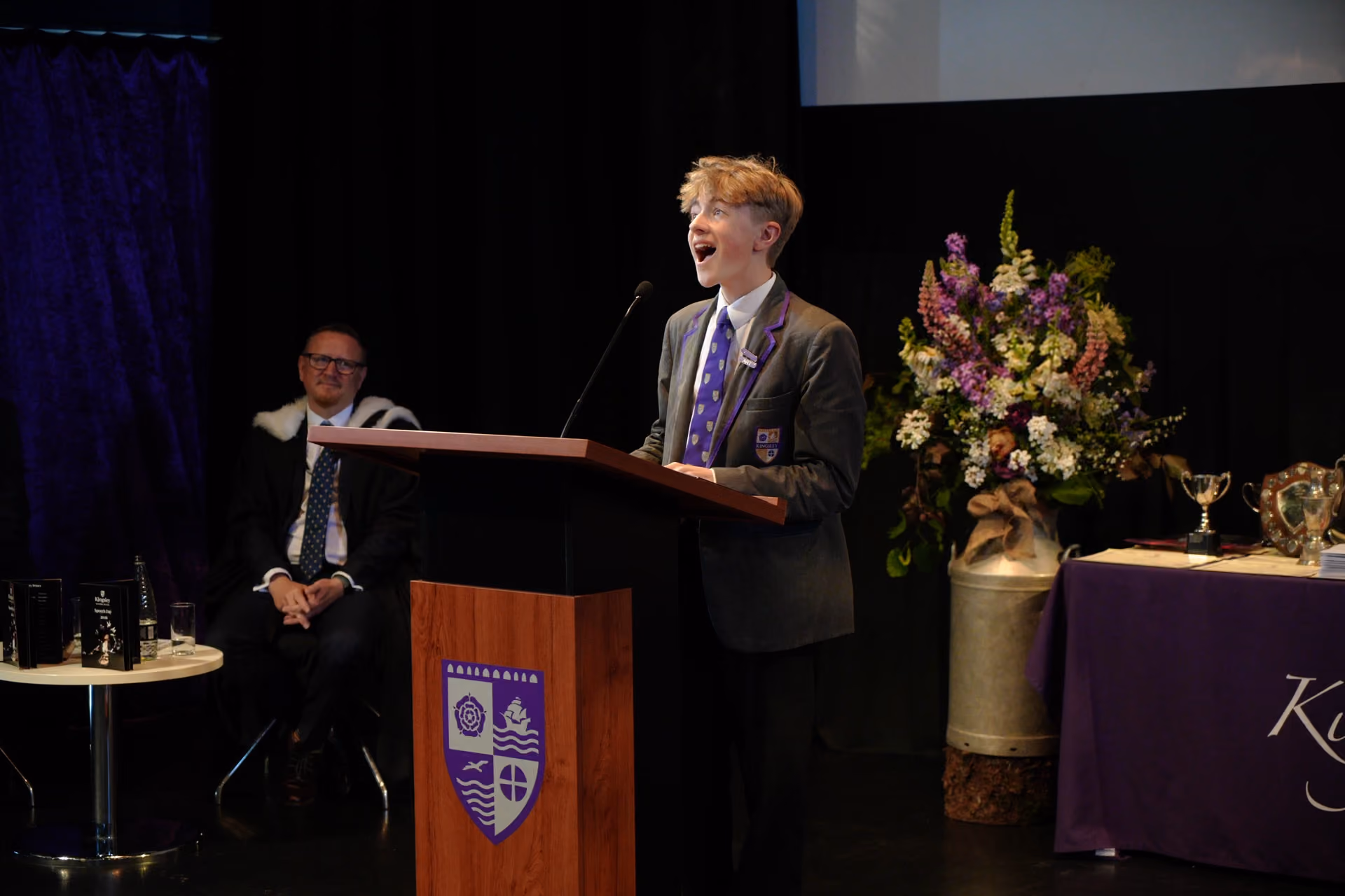 A young male student in a grey blazer and purple tie speaking enthusiastically at a wooden podium with a microphone during a school event, with a man seated in formal attire in the background and a table with trophies and flowers to the right.