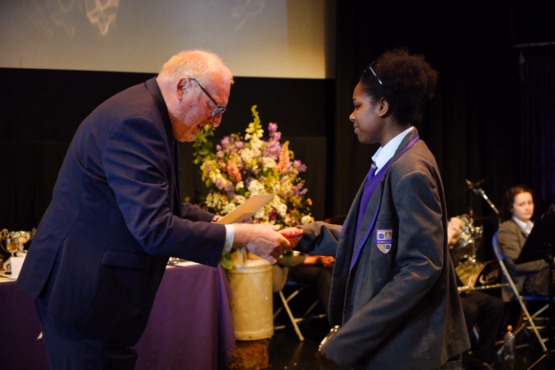 A man in a suit hands a certificate to a schoolgirl wearing a grey blazer with a purple sweater and school badge during an awards ceremony, with flower arrangements and seated students in the background.