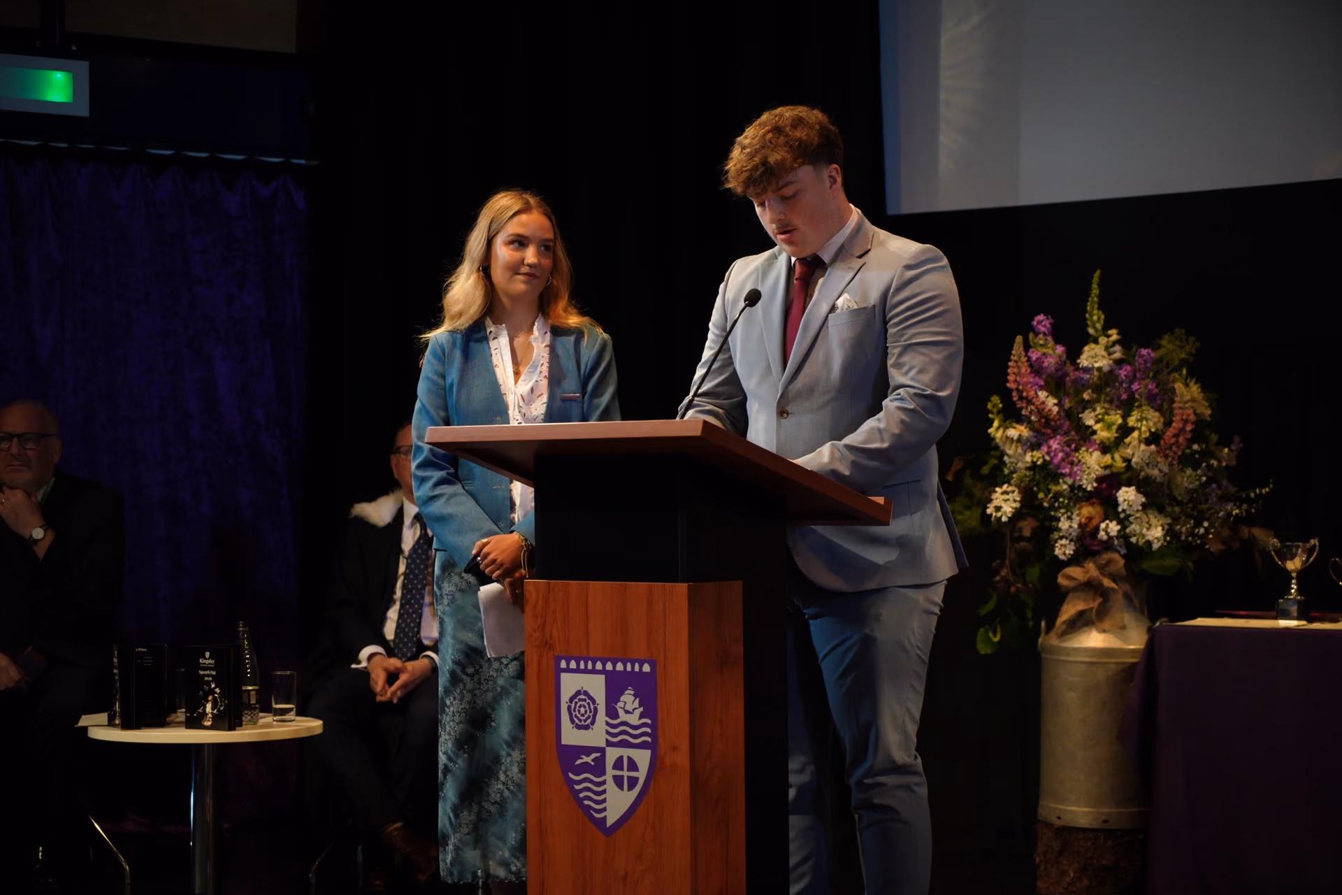 A young man in a grey suit and maroon tie is speaking at a wooden podium with a school crest, while a young woman in a teal blazer and floral skirt stands beside him, smiling. They are on a stage with seated audience members partially visible in the background, along with a large floral arrangement and a table with trophies.