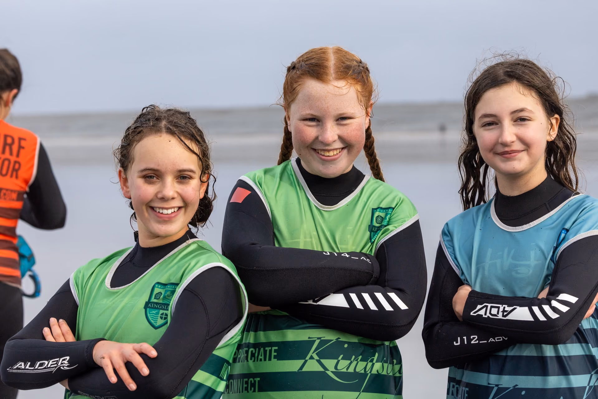 Three smiling young girls in wetsuits and green or blue surf vests standing with arms crossed on a beach with calm sea in the background.