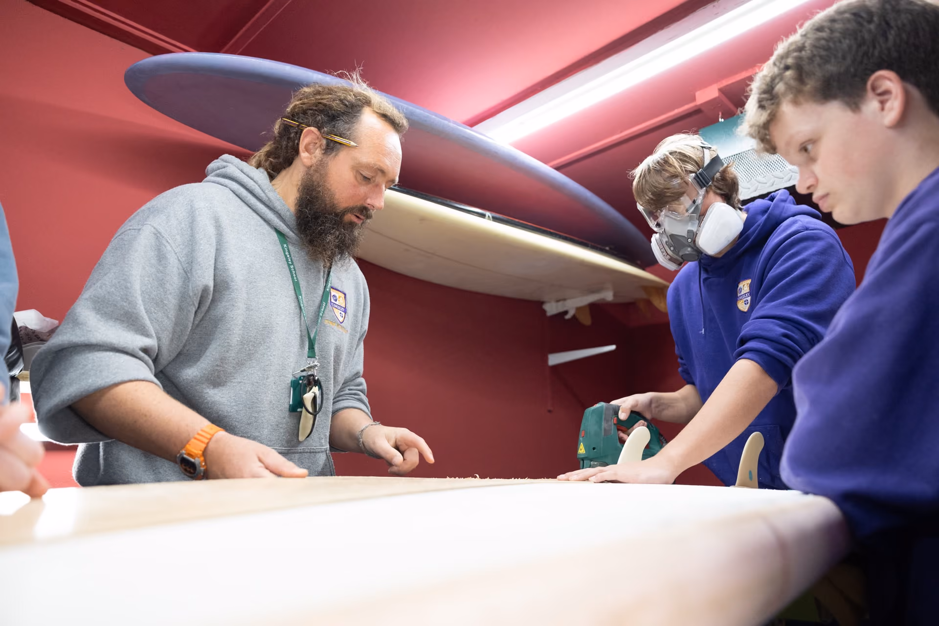 Three people in a workshop shaping a surfboard; one wears a respirator mask while sanding, another with a pencil behind his ear closely observes.
