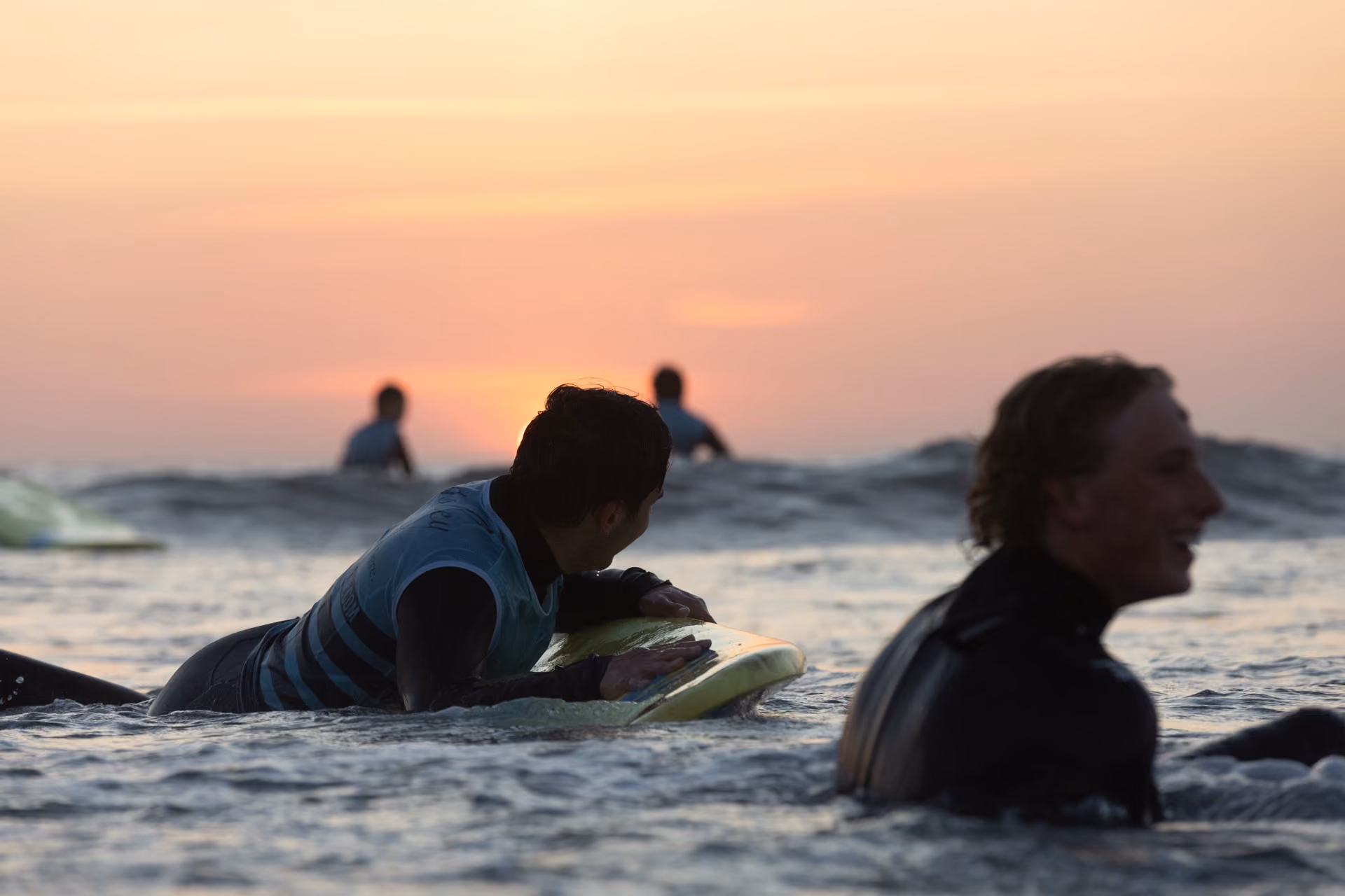 Two surfers in wetsuits paddling on surfboards in the ocean at sunset, with two more surfers visible in the distance near waves.