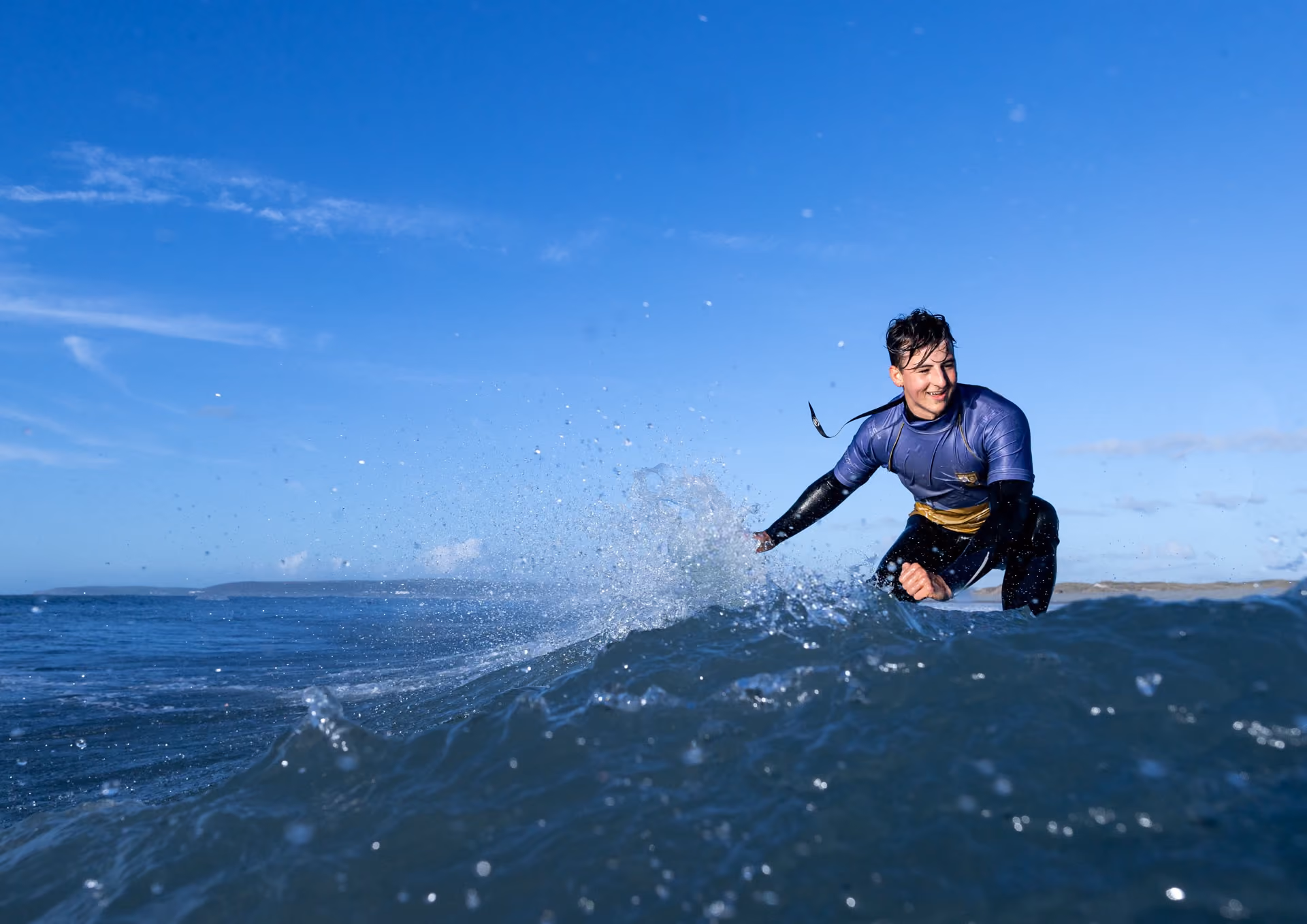 Surfer in a blue and black wetsuit riding a wave in the ocean under a clear blue sky, smiling and leaning forward.