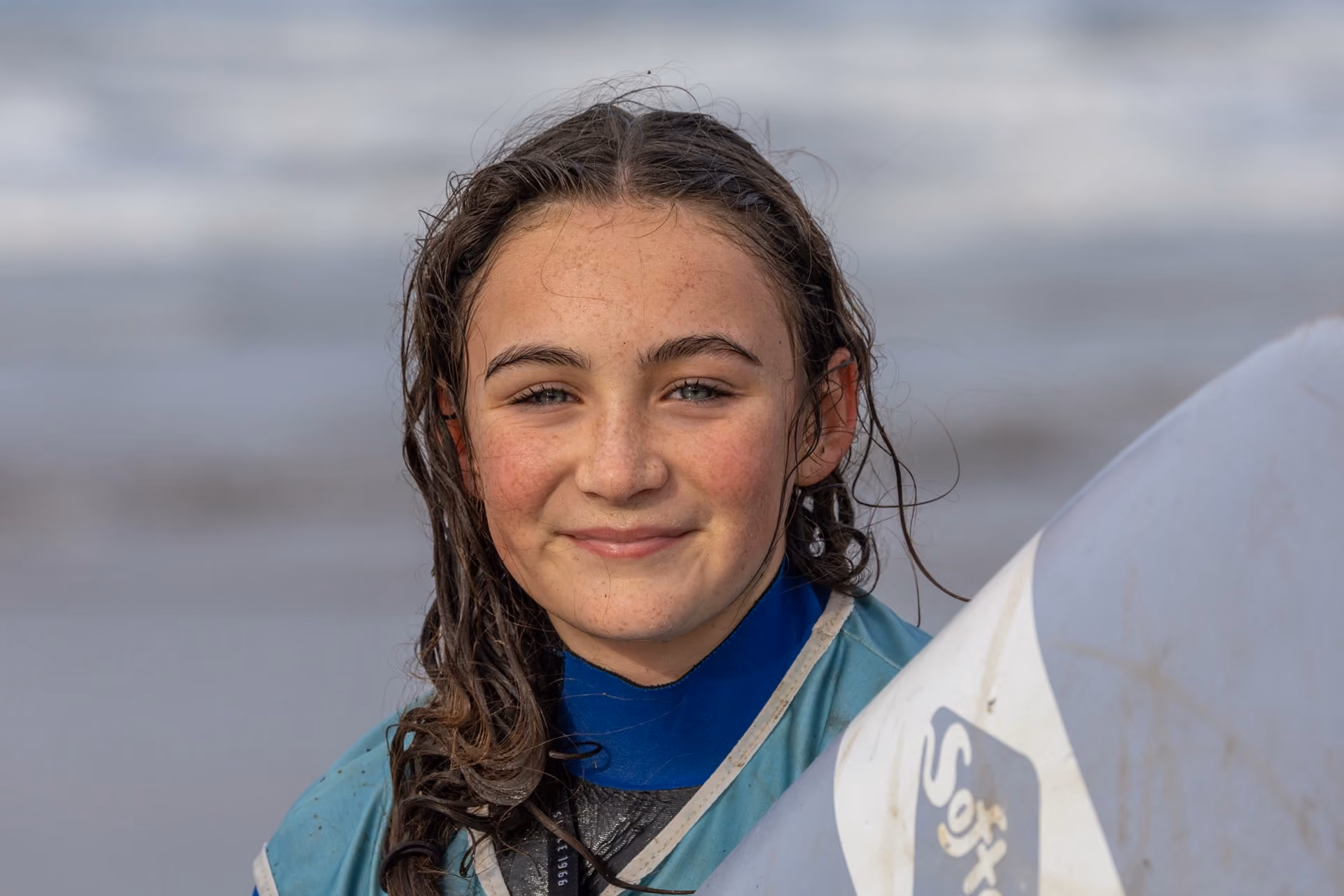 Portrait of a smiling young girl with wet hair in a blue wetsuit holding a white surfboard at the beach.
