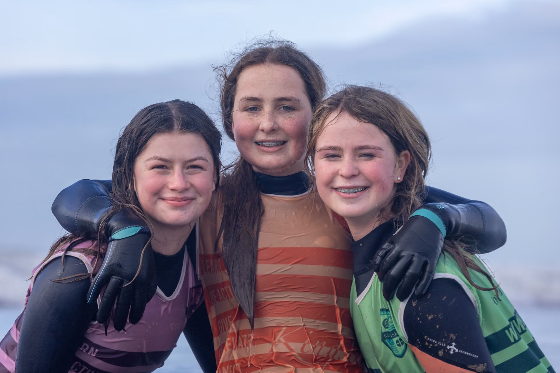 Three smiling teenage girls with wet hair wearing wetsuits and coloured vests embrace each other outdoors near water and mountains.