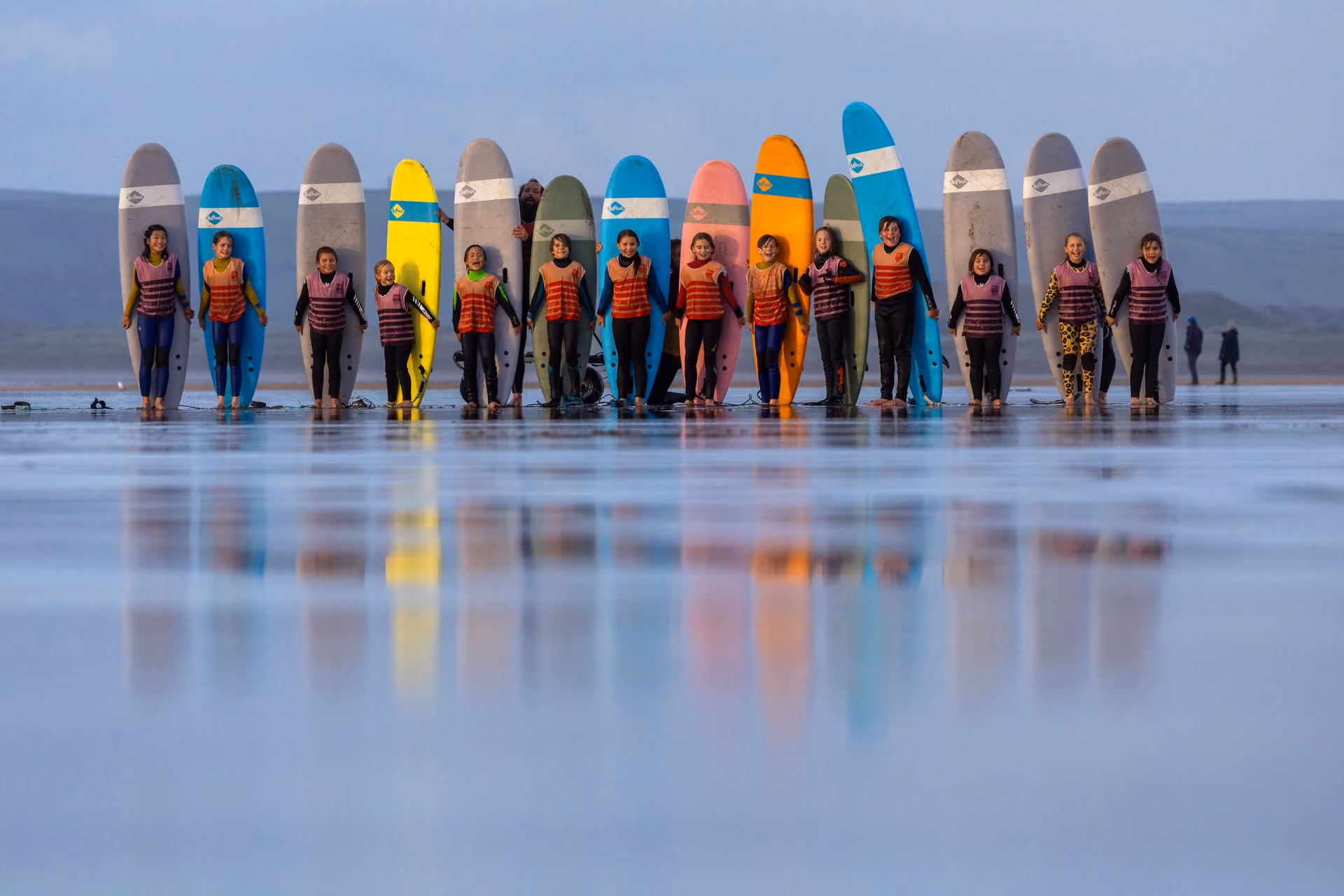 A group of children standing in a line on a beach holding colorful surfboards upright behind them, wearing wetsuits and orange or pink life vests, with their reflections visible on the wet sand.
