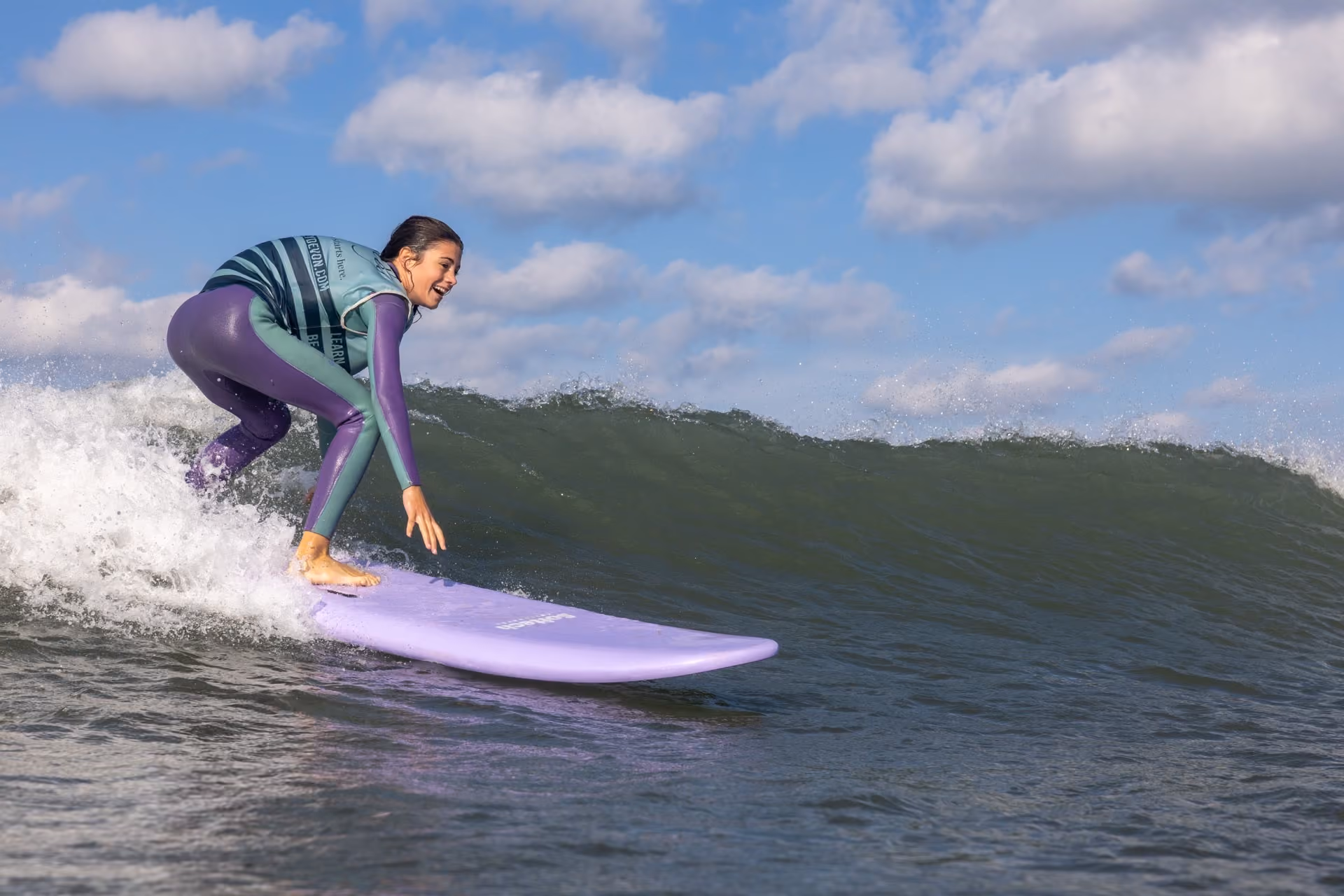 Woman in a wetsuit riding a purple surfboard on a breaking wave under a partly cloudy sky.