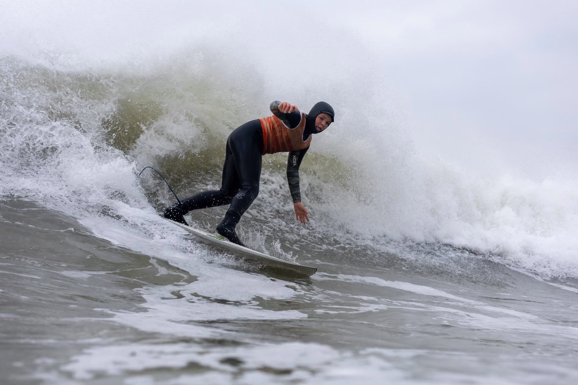 Surfer wearing a black wetsuit and orange vest riding a wave in misty conditions with water splashing around.