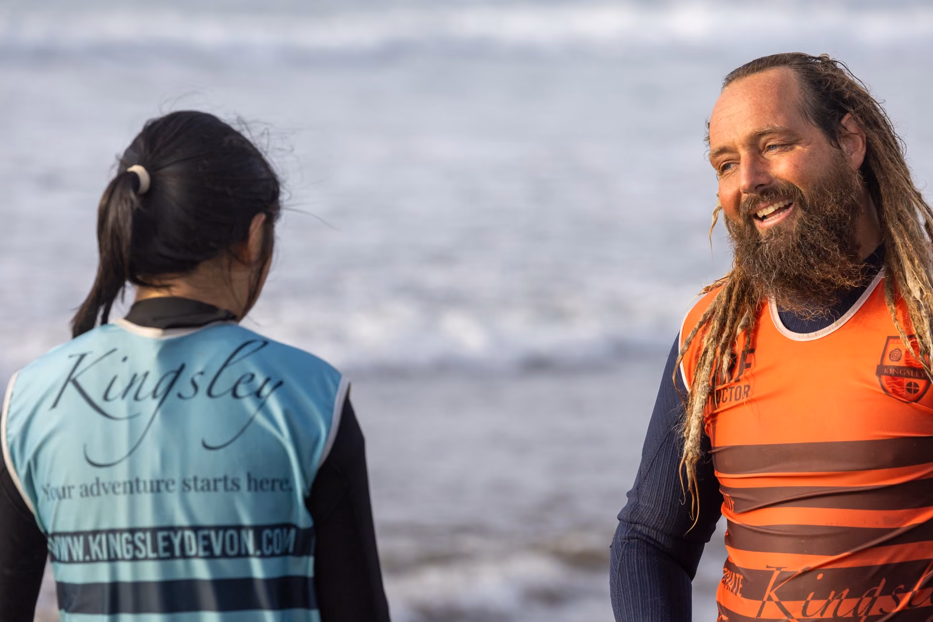Two people in wetsuits standing by the sea, one with long dreadlocks and beard wearing an orange vest smiling at a woman with a ponytail and blue vest with Kingsley branding on the back.