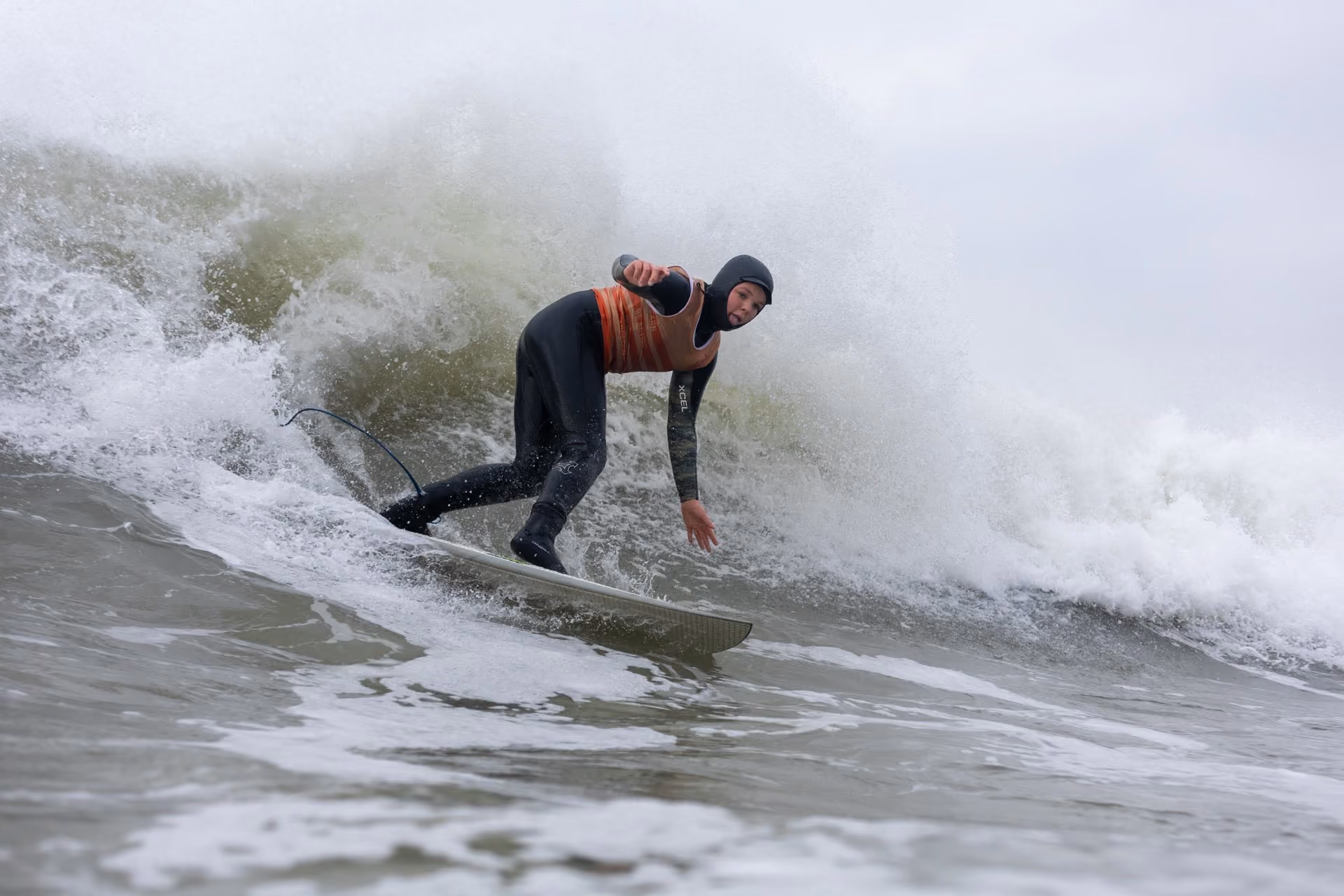 Surfer in a black wetsuit and orange vest rides a large wave in cloudy conditions.