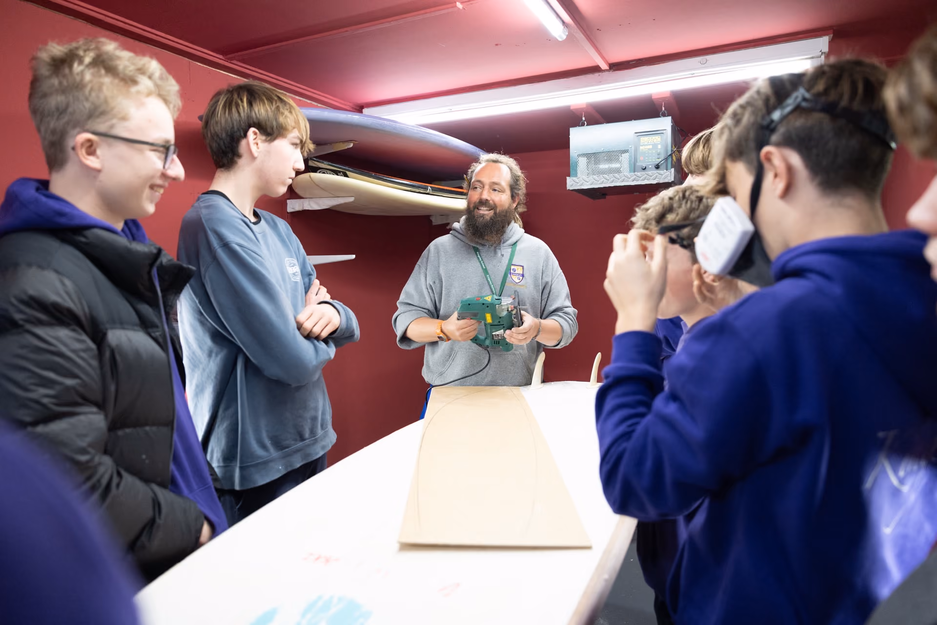A bearded instructor in a grey hoodie demonstrating a tool on a wooden board to a group of attentive young students inside a workshop with maroon walls and surfboards mounted on the wall.